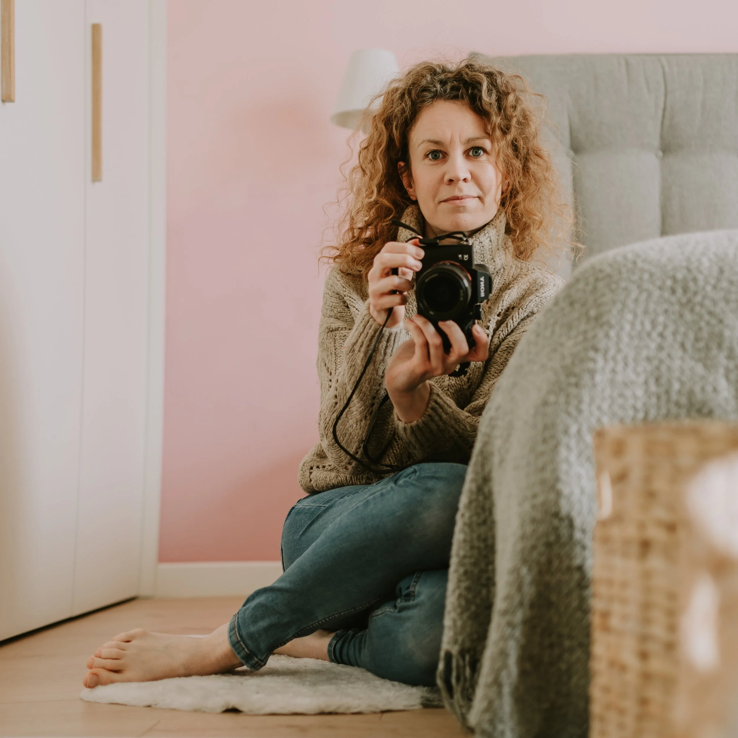 A woman with curly hair taking a photo with a camera while sitting on the floor in a bedroom with pink walls, gray headboard, and beige blankets.