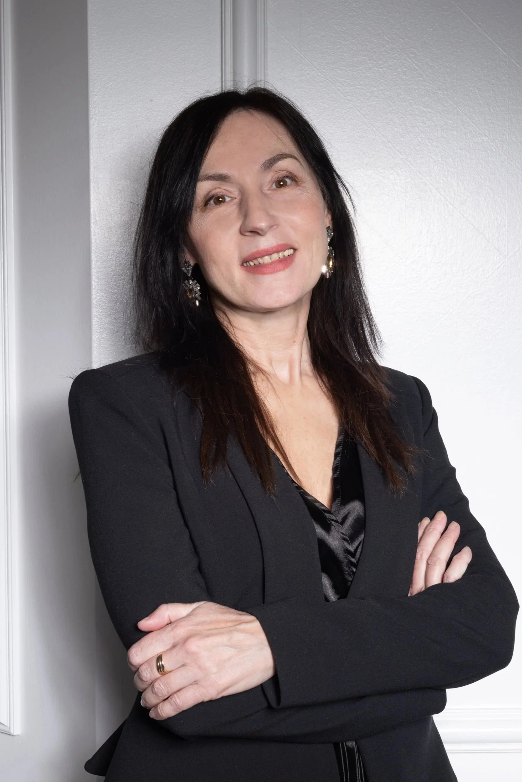 A woman with black hair, wearing a black blazer and earrings, standing with arms crossed against a white wall.