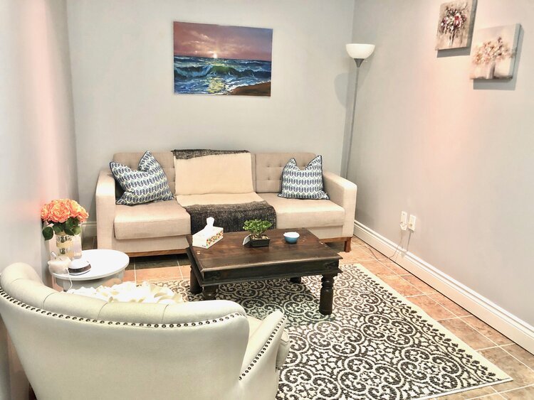 Living room with a beige sofa, two patterned pillows, a dark wood coffee table, an armchair, a side table with a vase of pink flowers, a patterned rug, a tall floor lamp, and wall art including a seascape and floral paintings.