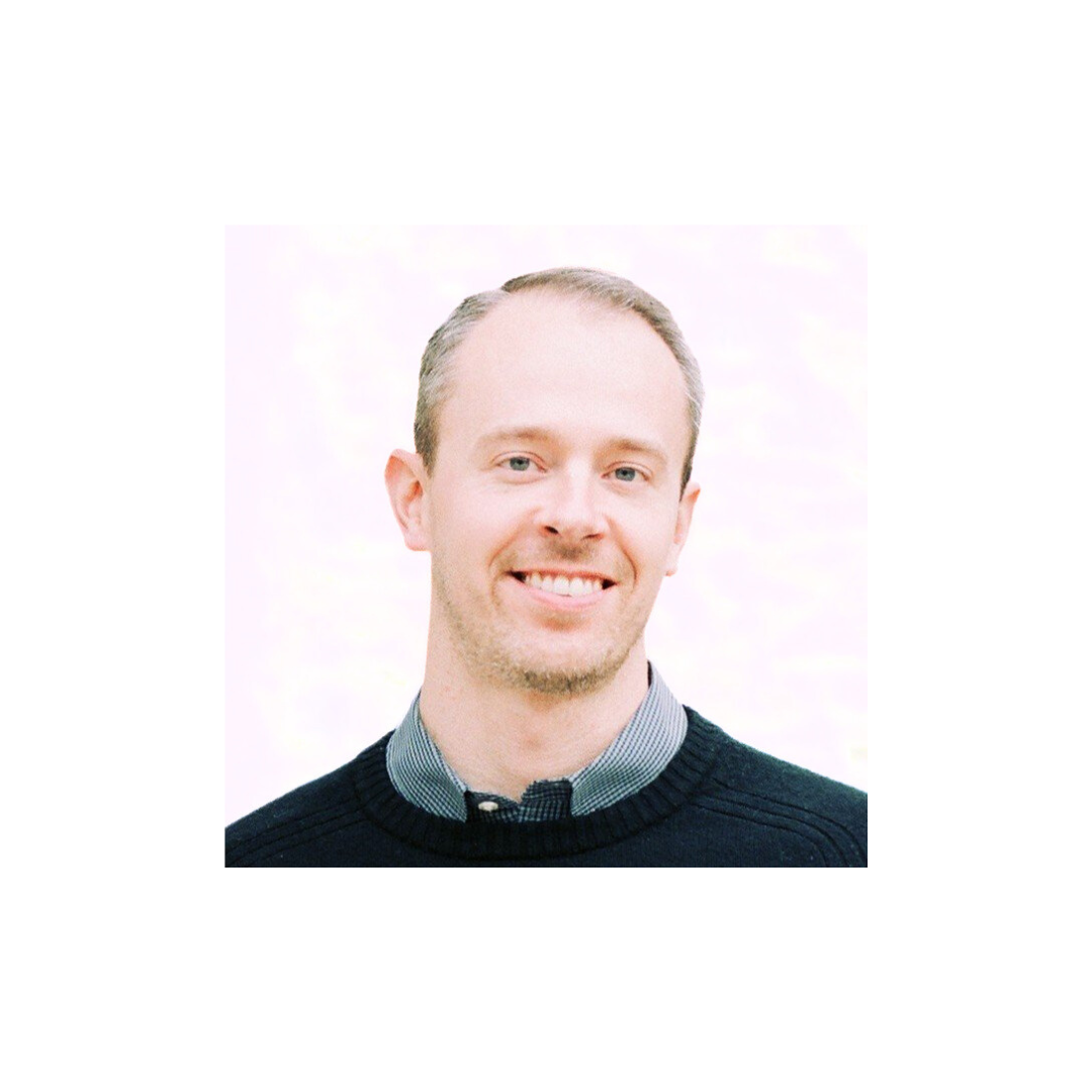 Headshot of a man with short light-colored hair, smiling, wearing a dark sweater over a gray collared shirt.
