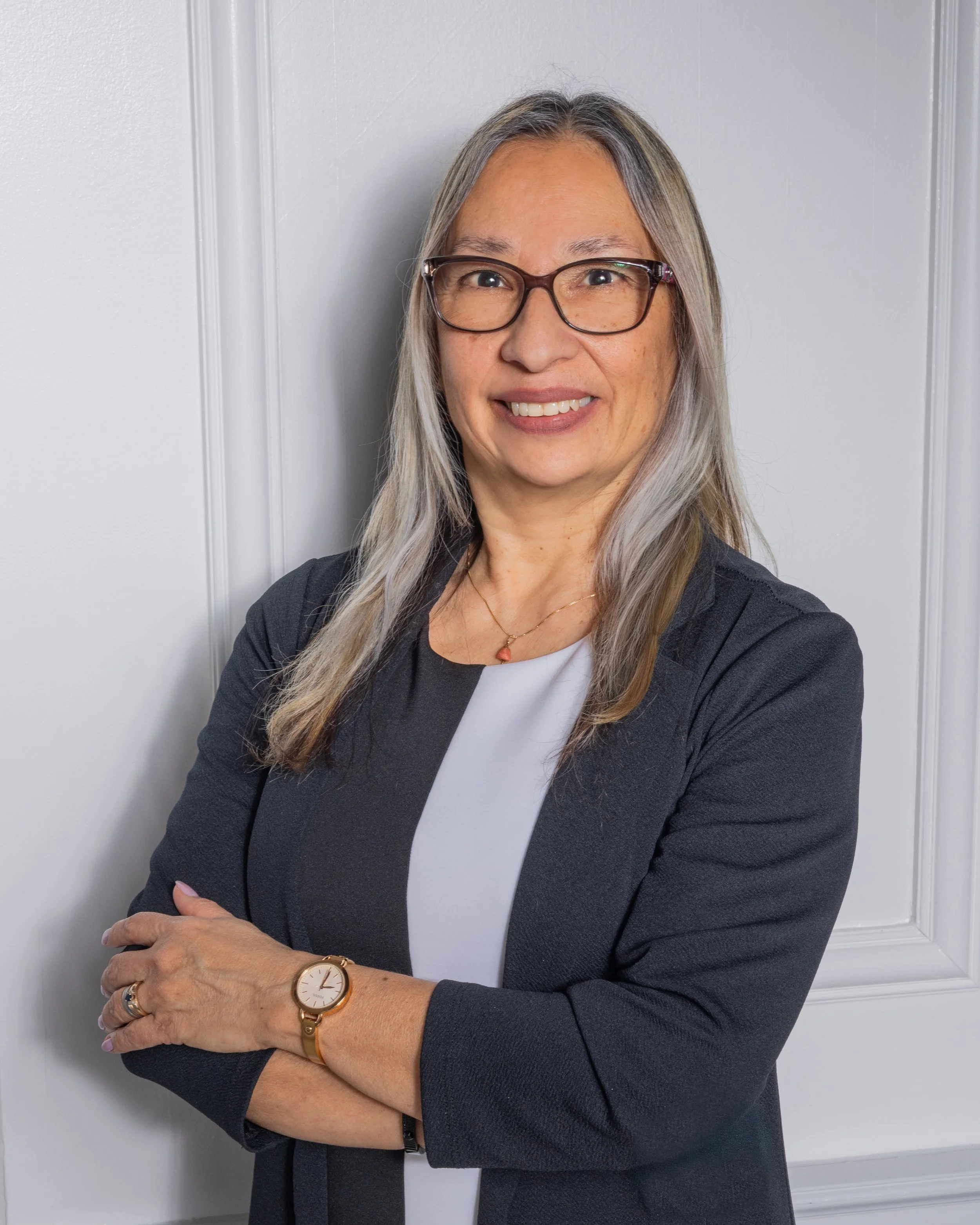 A woman with glasses and long gray hair smiling, standing with arms crossed in front of a white wall, wearing a black blazer over a white top, with a watch and necklace.