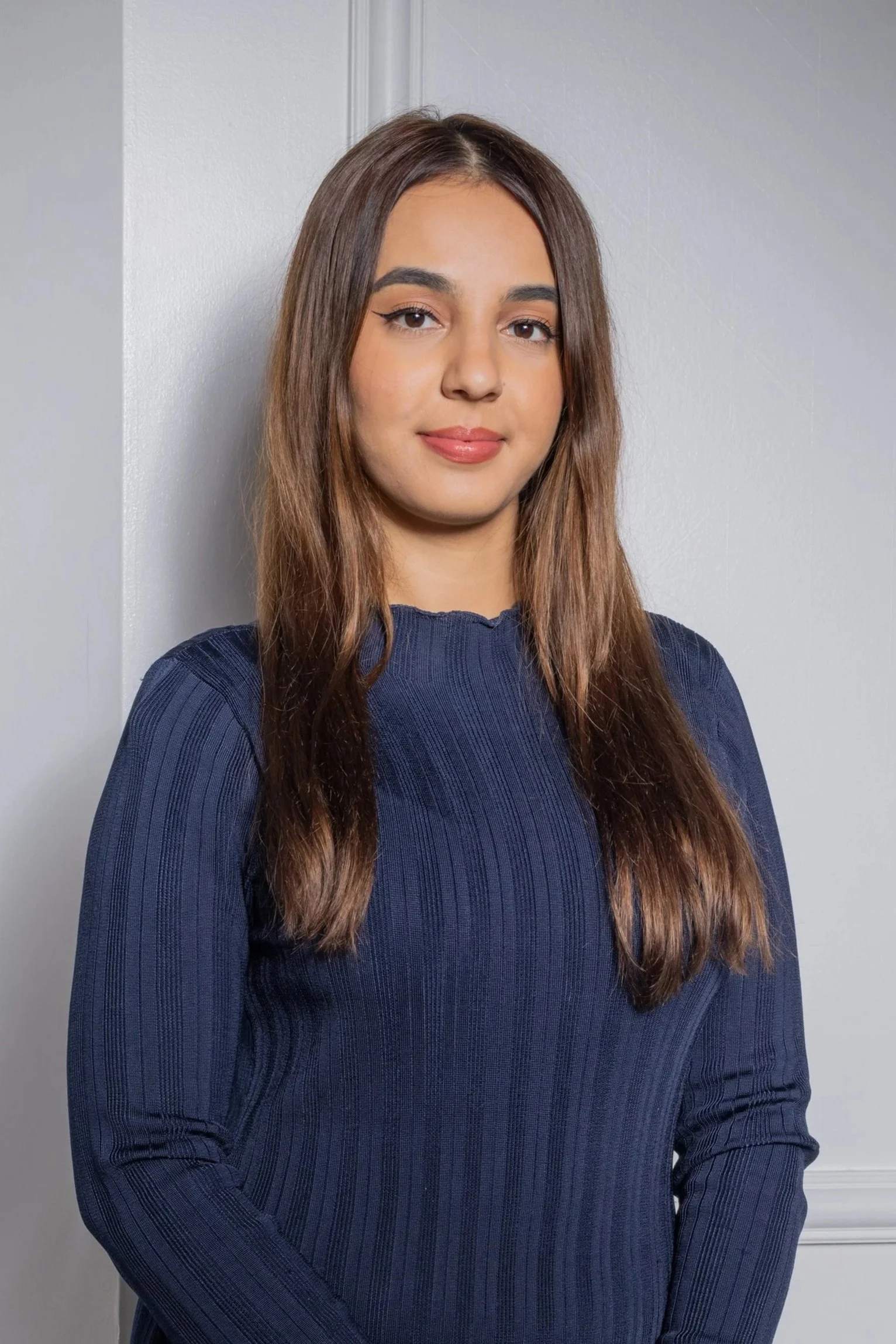 A young woman with long brown hair, wearing a navy blue ribbed sweater, standing against a white wall with a doorframe.