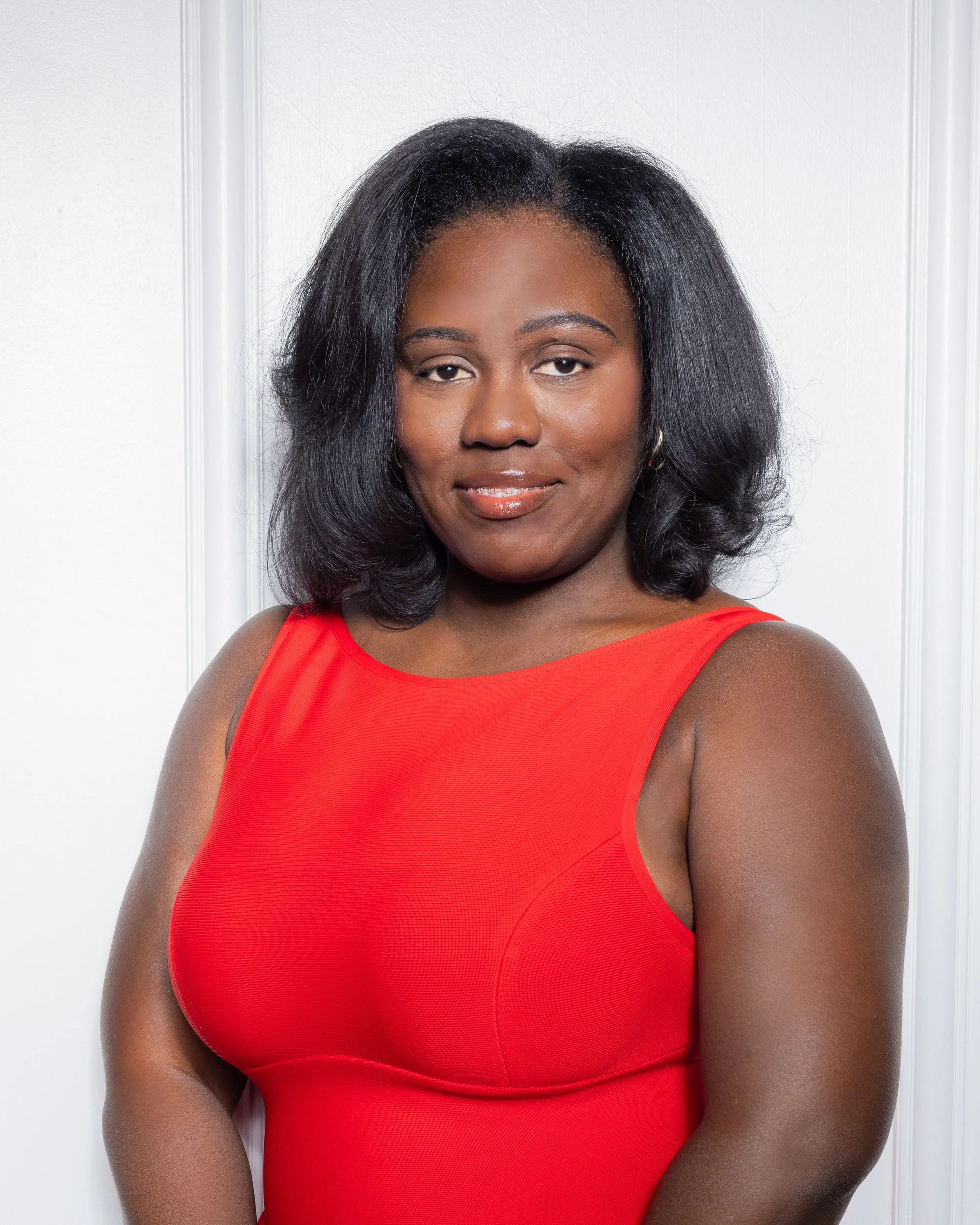 A woman with shoulder-length black hair wearing a red sleeveless dress, standing against a white wall with decorative molding.