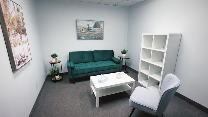 A small waiting room with a green velvet sofa, a white coffee table, a white bookshelf, an upholstered armchair, and framed artwork on the walls.