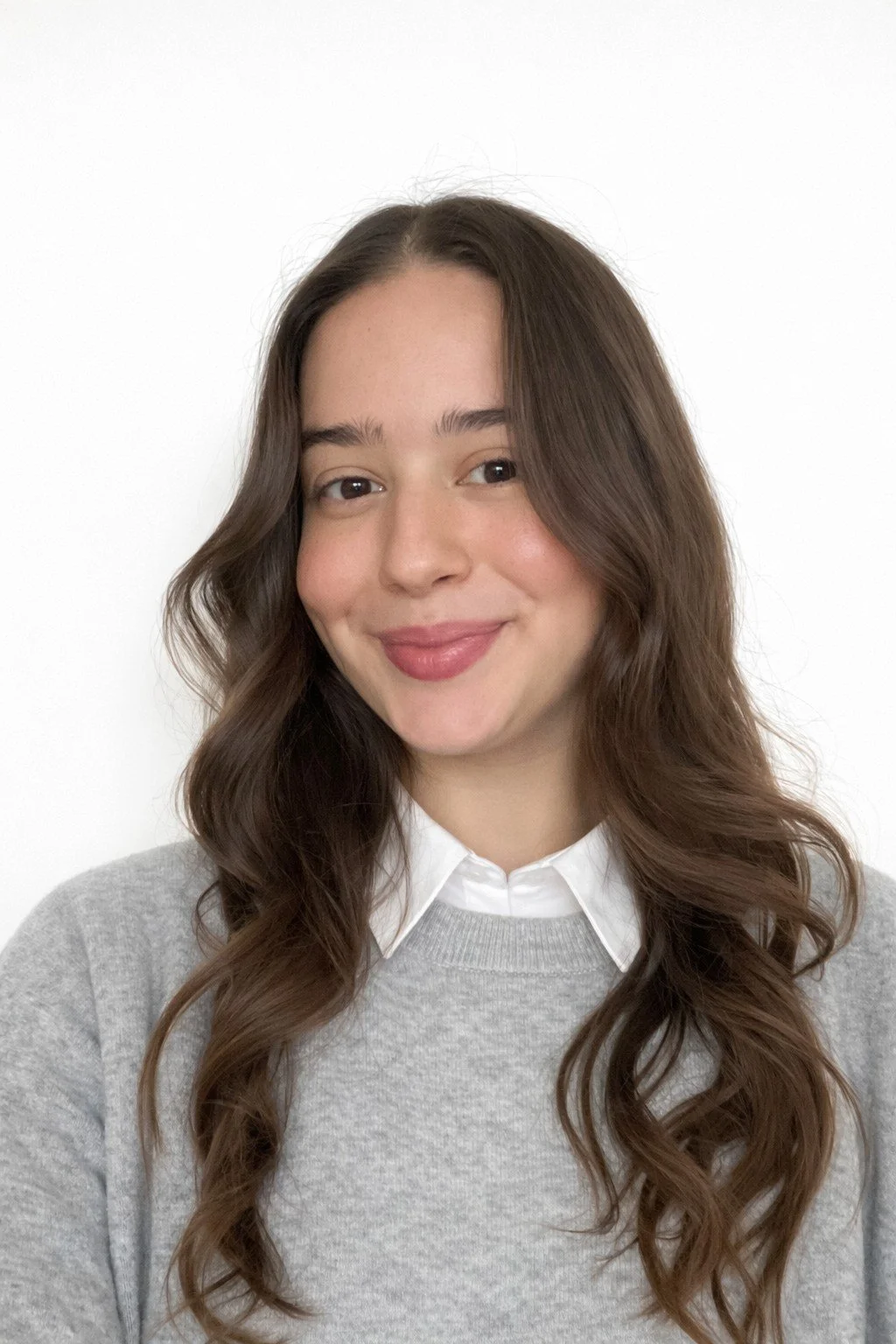 Portrait of a young woman with long, wavy brown hair, smiling, wearing a white collared shirt under a light gray sweater, against a plain white background.