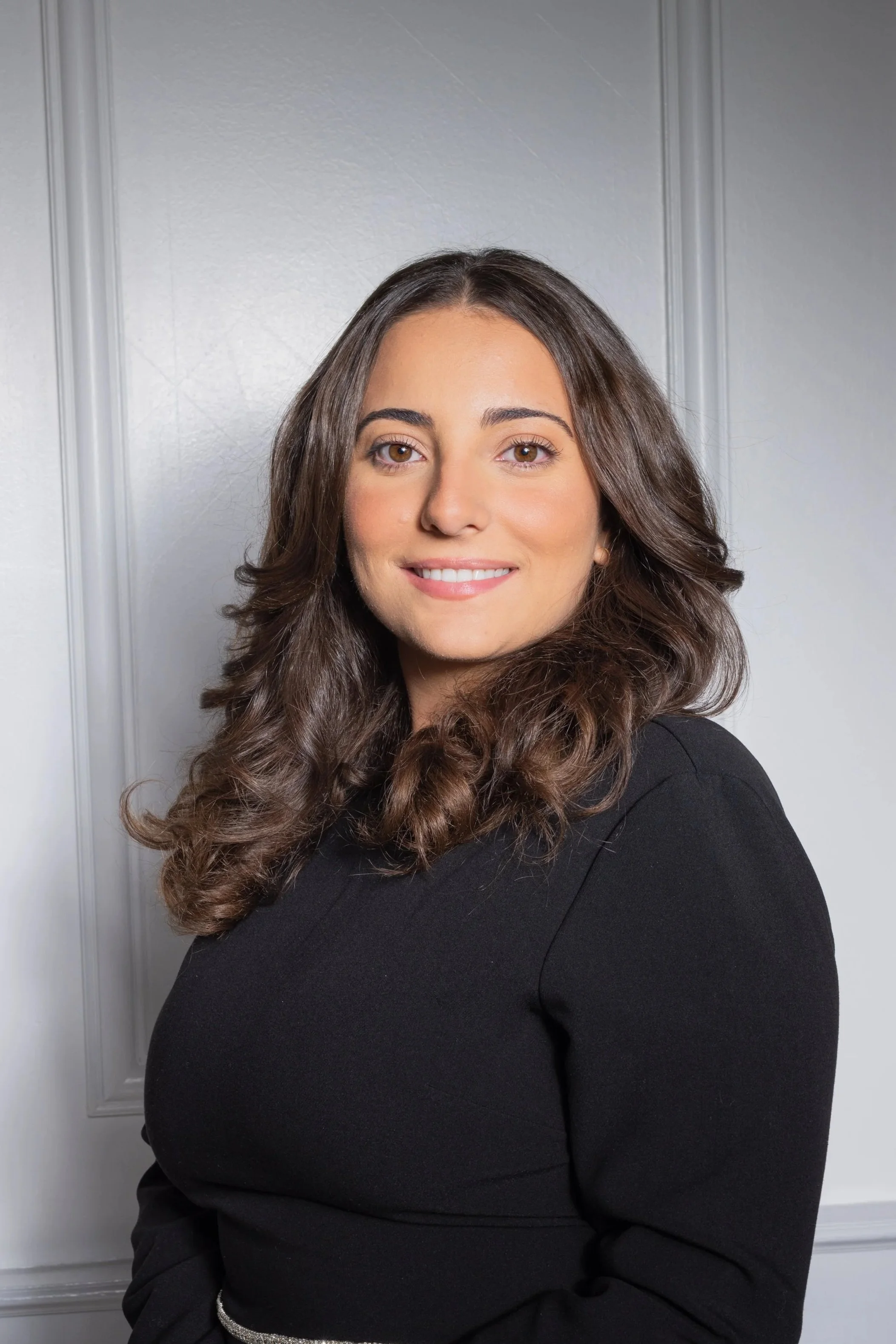 A portrait of a smiling woman with wavy brown hair wearing a black top, standing against a light gray wall with decorative paneling.