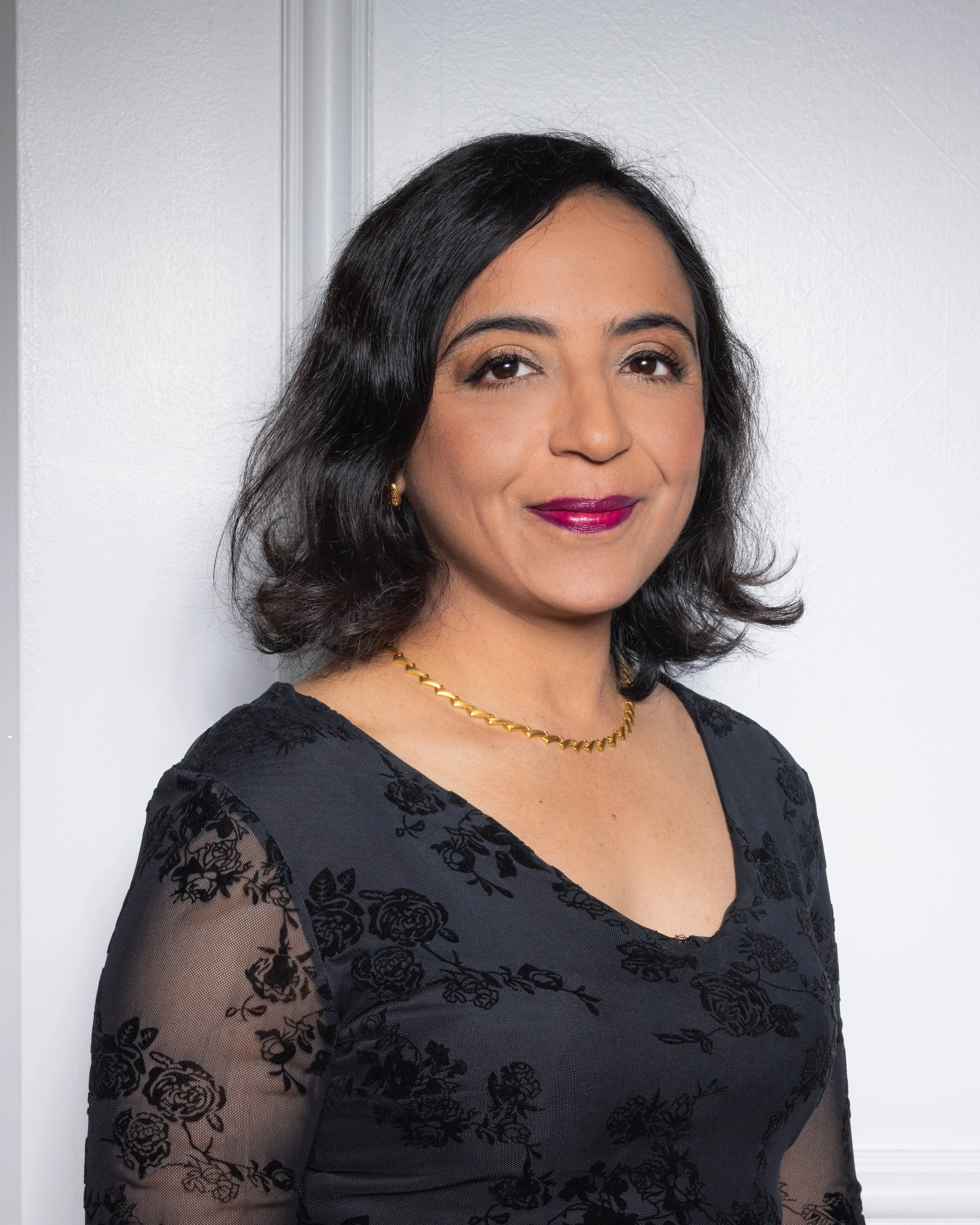 Portrait of a woman with dark hair, wearing a black floral lace top with transparent sleeves, accessorized with gold jewelry, standing against a white wall.