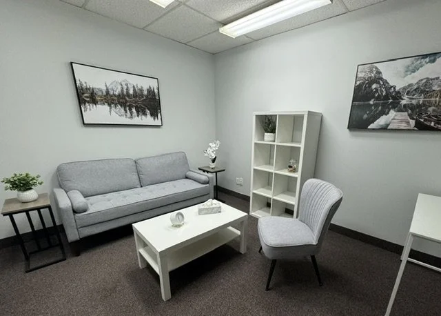 A waiting room with a gray sofa, a small white coffee table, a gray upholstered chair, a white bookshelf, a side table with a plant, and framed landscape photos on the walls.
