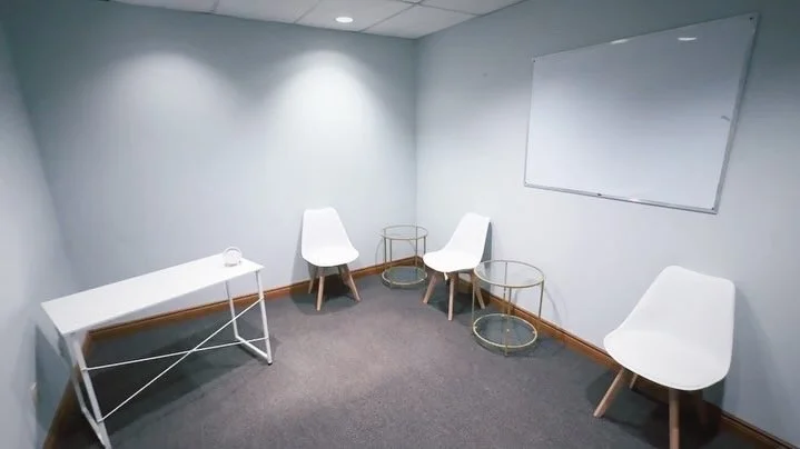 Empty room with four white chairs, a white table, gold and glass side tables, whiteboard on the wall, and gray carpet.