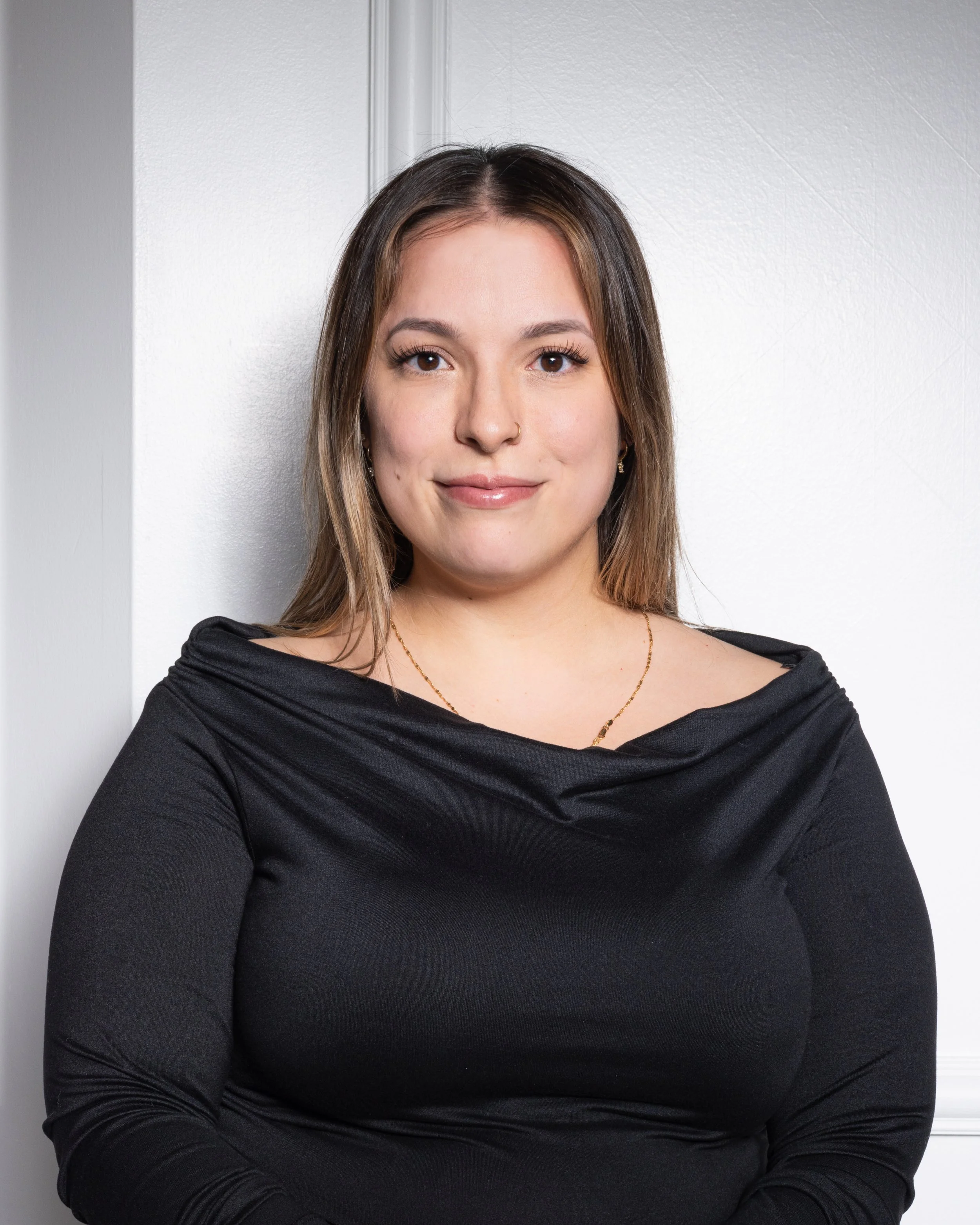 Portrait of a young woman with light brown hair, wearing a black top, standing against a white wall.