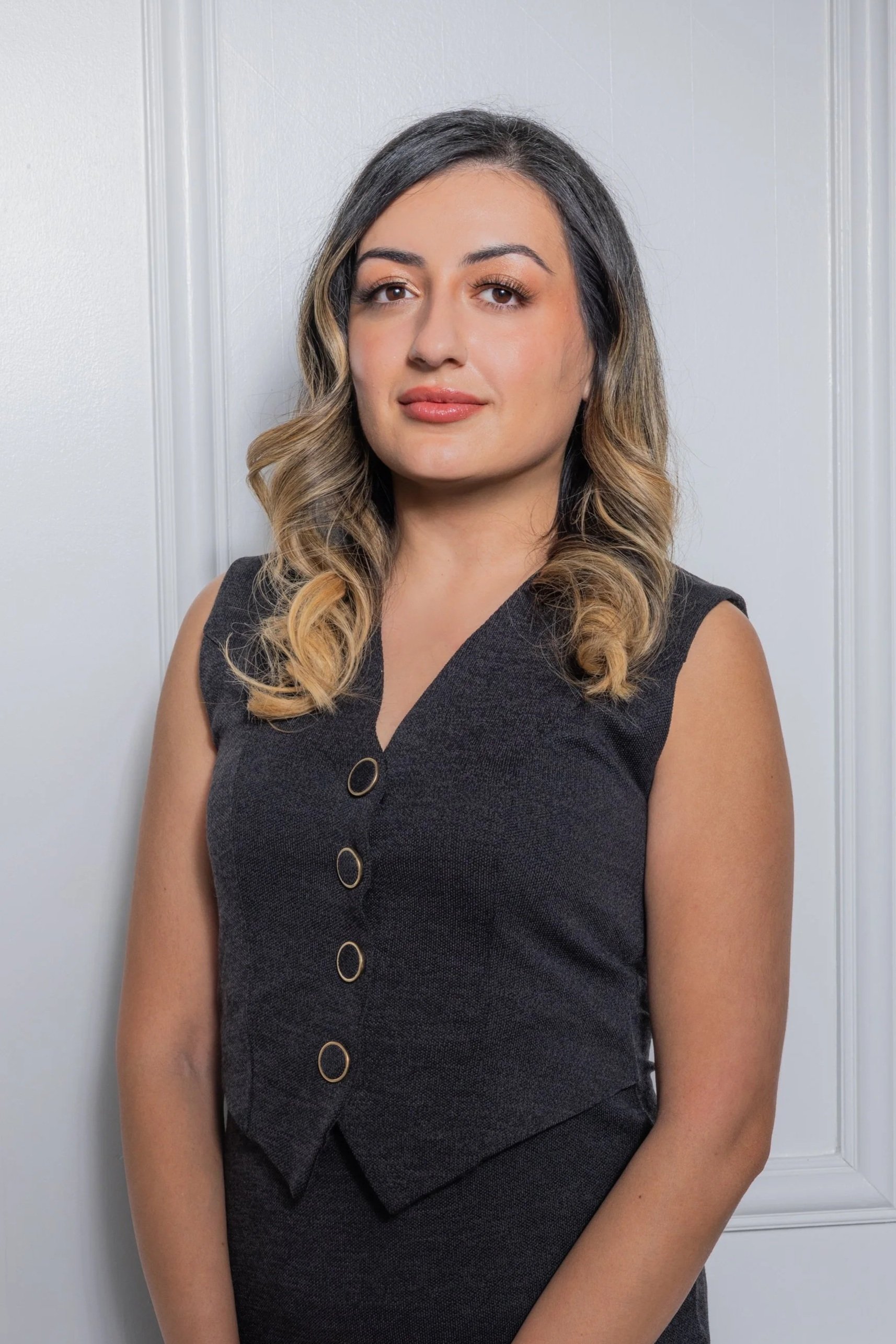 A woman with shoulder-length wavy hair styled in loose curls, wearing a sleeveless black dress with a v-neckline and decorative buttons, posing against a light-colored wall with white paneling.