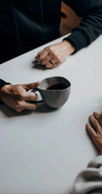 Person holding a dark coffee mug on a white table, with another person sitting nearby.