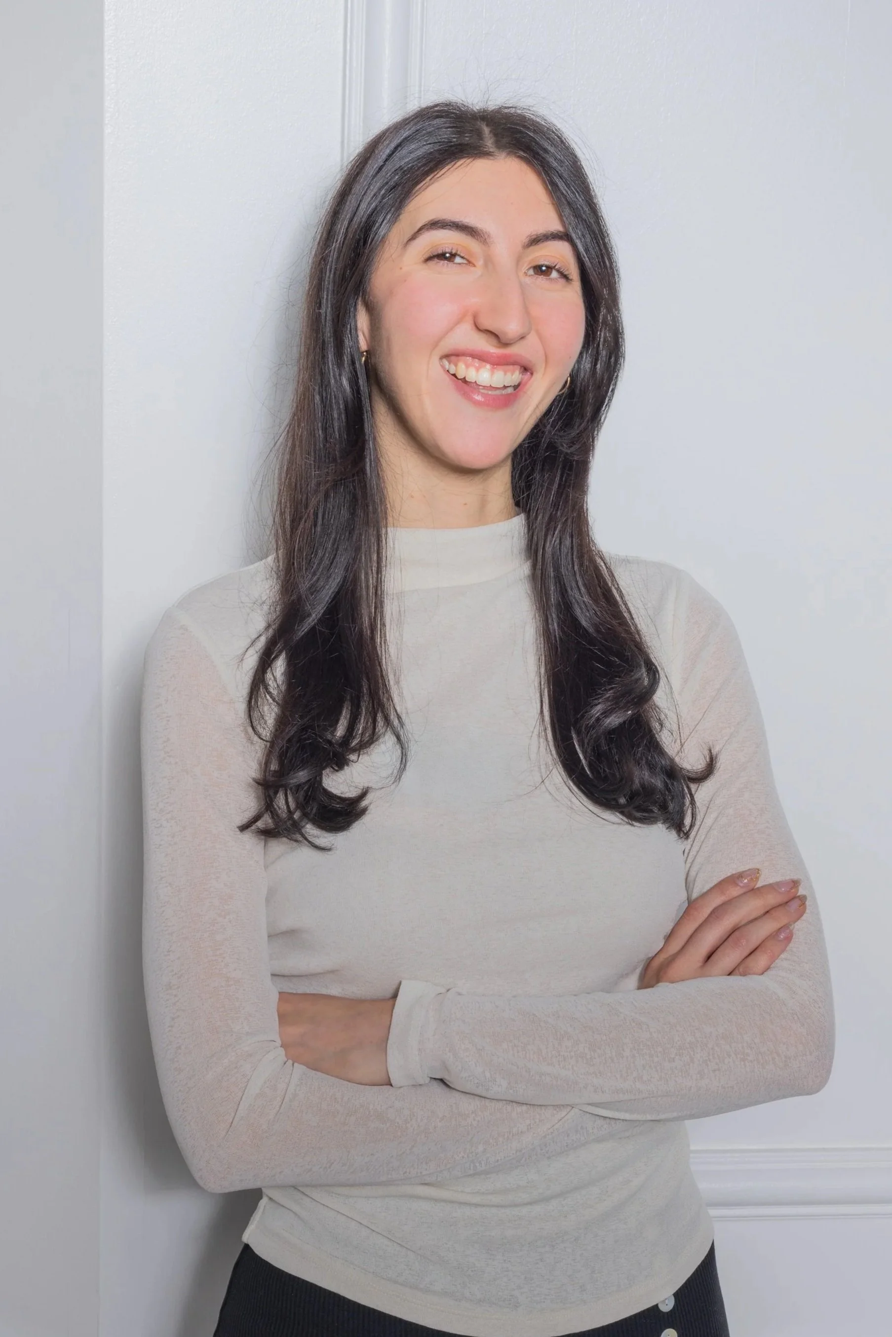 A woman with long dark hair, light skin, and a friendly expression, standing with arms crossed against a white wall, wearing a light-colored long-sleeve top.