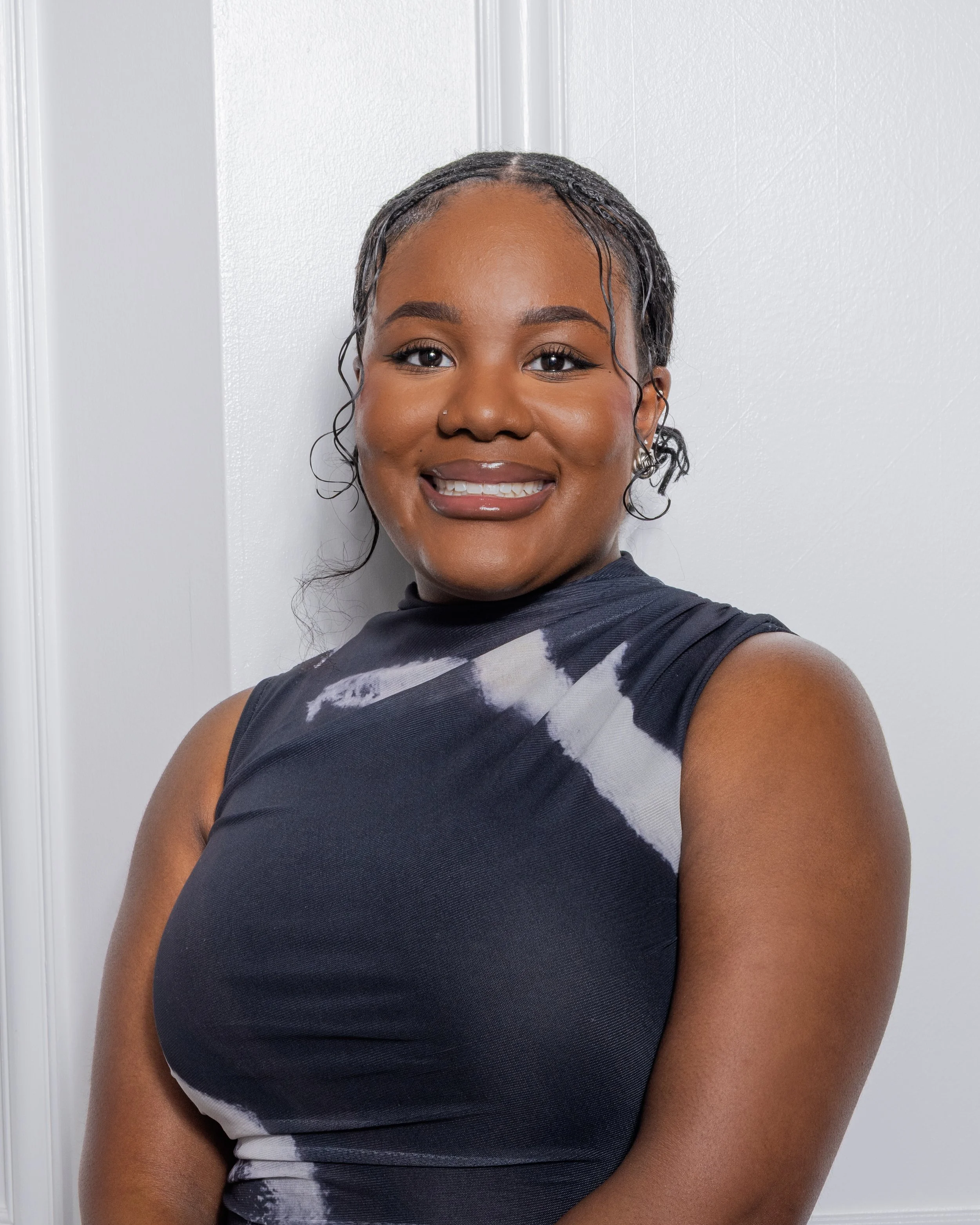 A young woman with dark skin, braided hair, and wearing a sleeveless black and gray tie-dye top, smiling while standing against a white door.