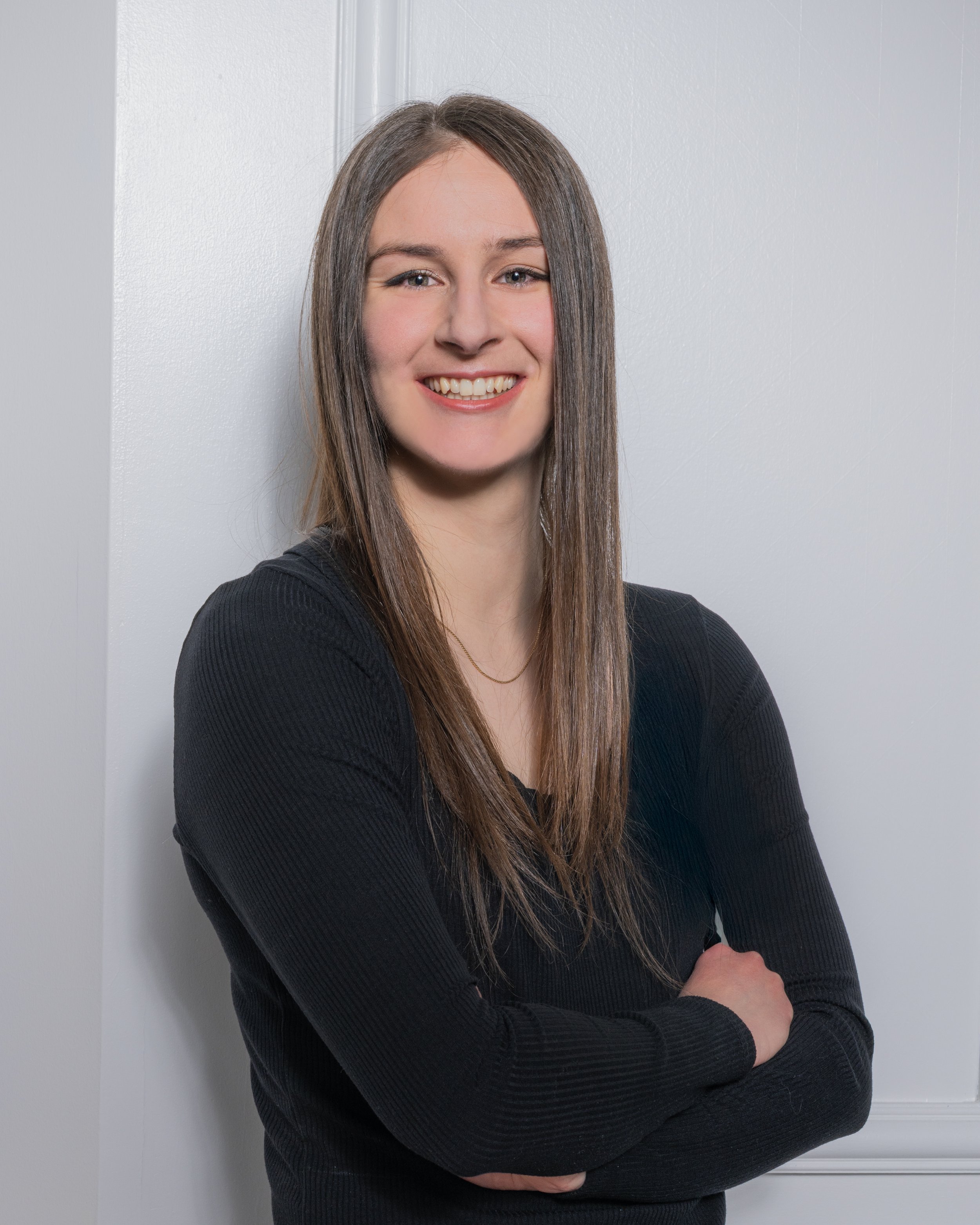 A young woman with long brown hair, smiling, wearing a black long sleeve top, standing against a white wall with her arms crossed.
