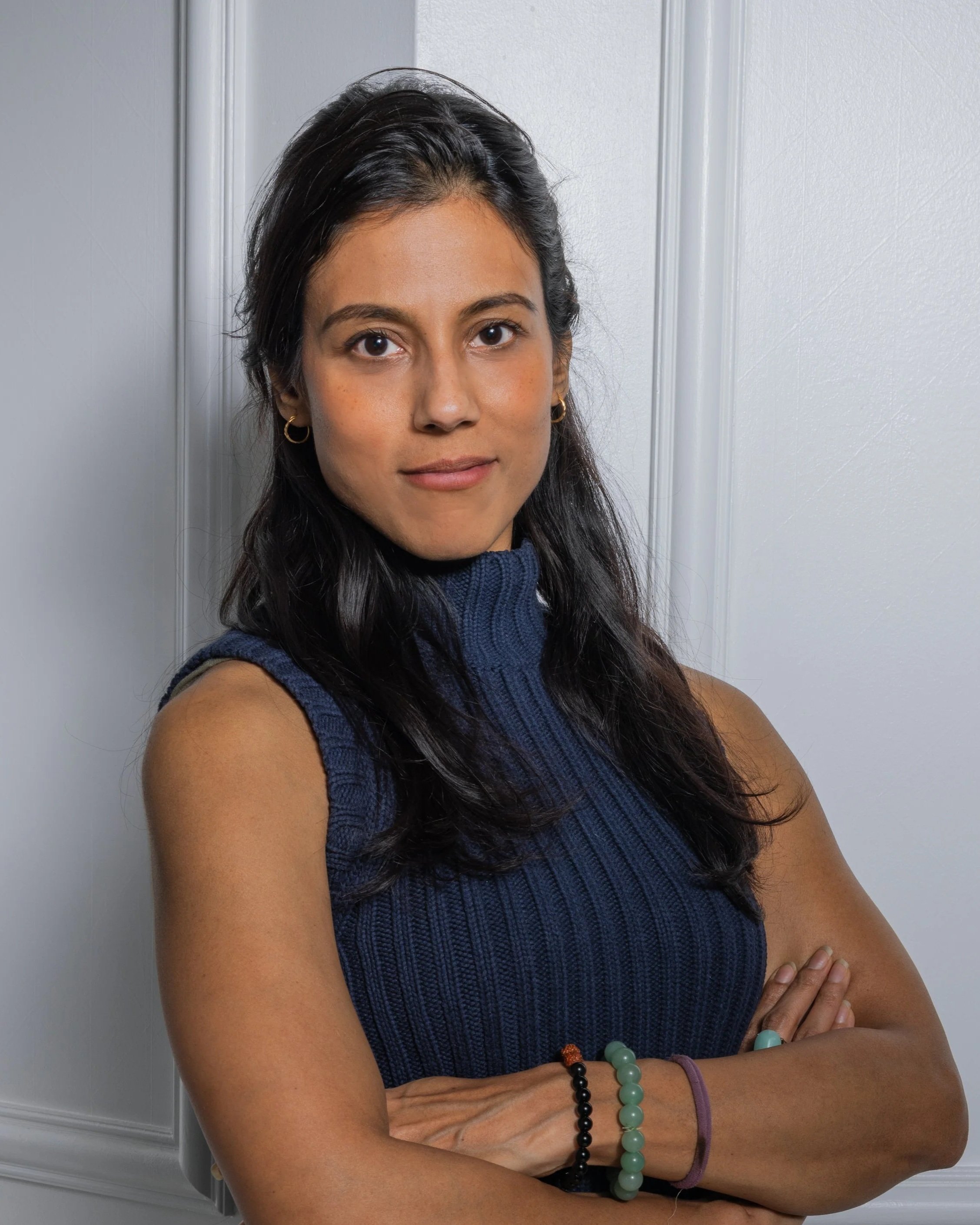 A woman with dark hair wearing a sleeveless blue knit turtleneck sweater, standing against a white wall, with arms crossed, looking at the camera.
