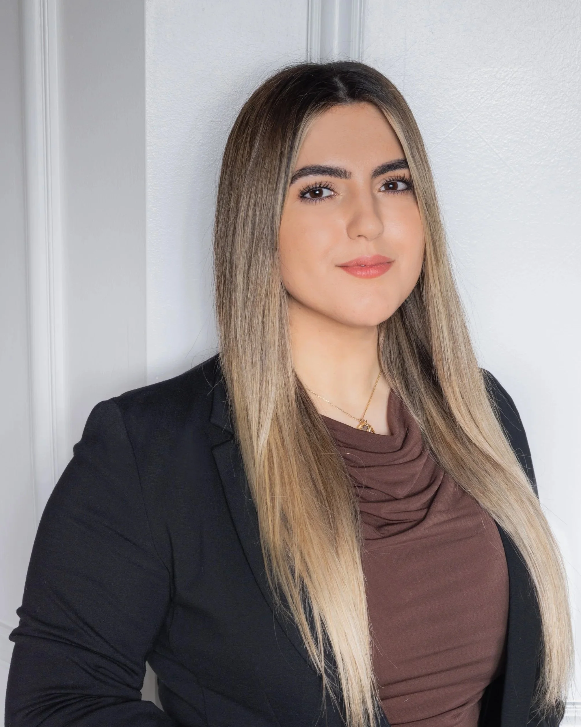 A woman with long blonde hair, wearing a black blazer over a brown top, standing against a white wall.
