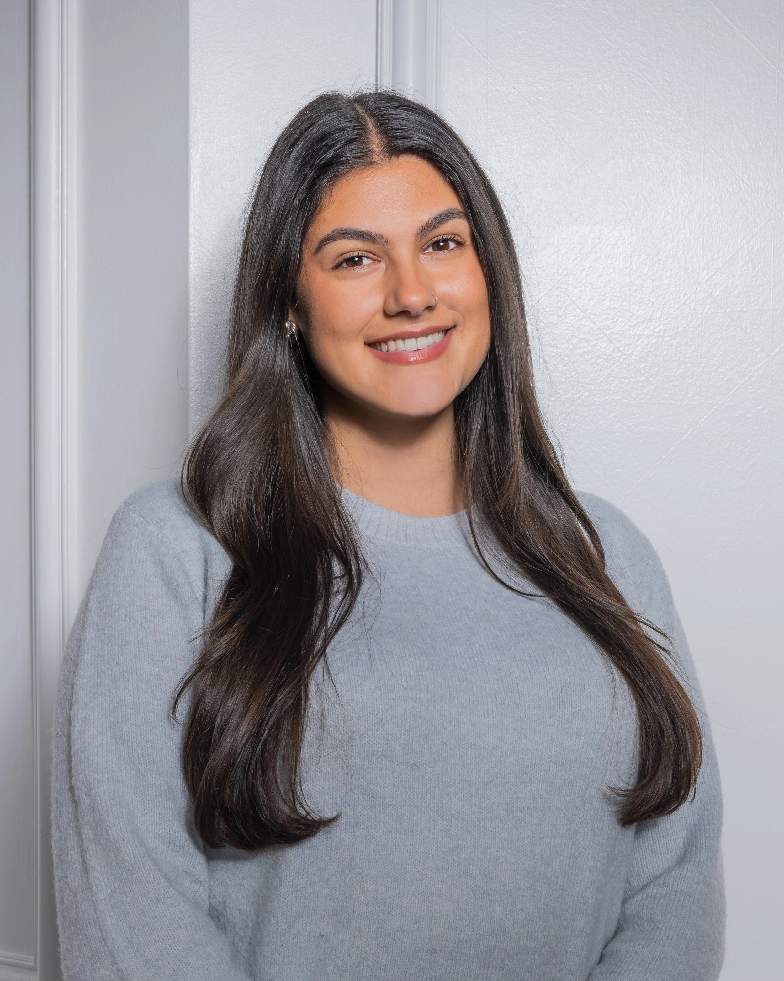 A young woman with long dark hair, smiling, wearing a light gray sweater, standing in front of a white wall with molding.