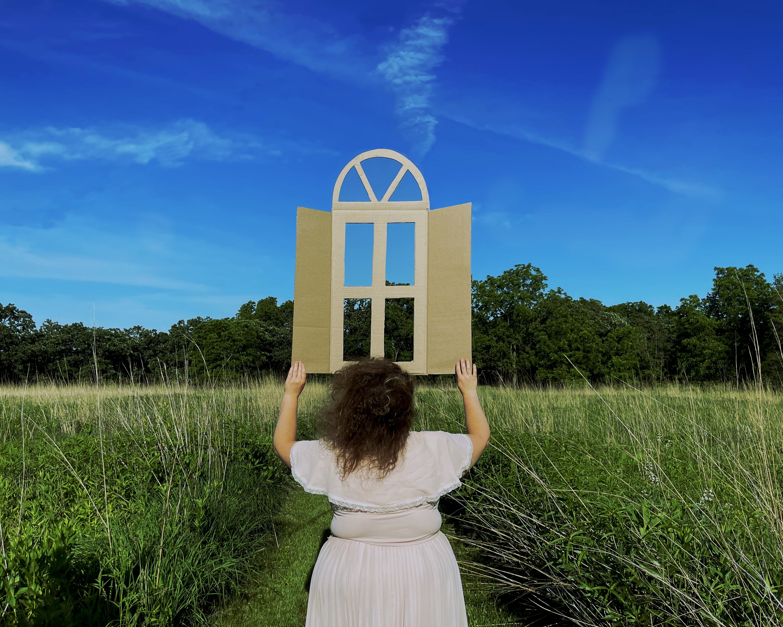A woman with wind-swept hair stands facing away from us in a prairie with tall grass under a bright blue sky. She opens a the shutters of a cardboard window that seems to float in front of her. Photo by Alex Benito Rodriguez with Katie Mazzini.