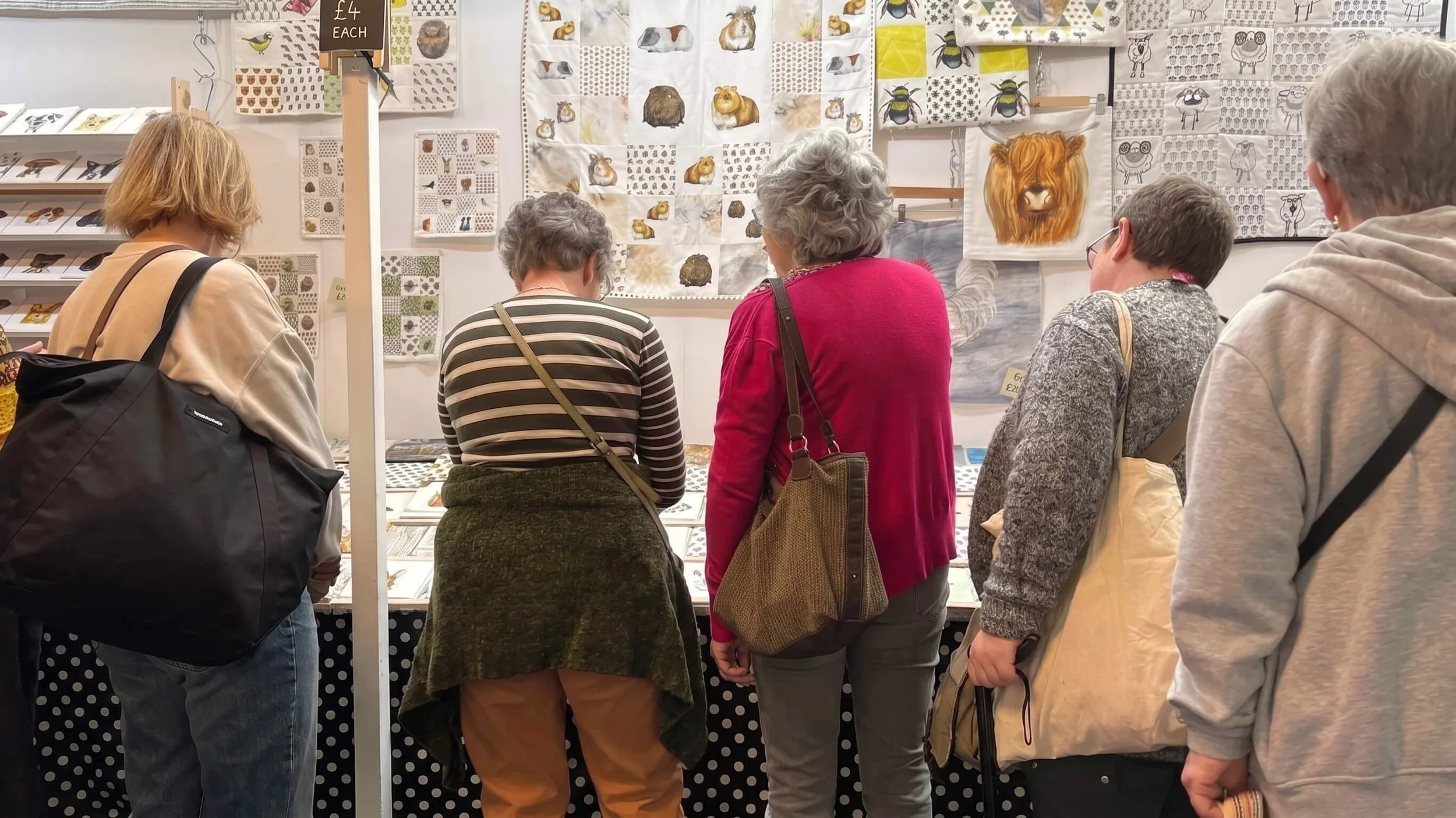 A group of five people standing at a booth looking at fabric or textile displays with animal patterns, including bears, guinea pigs, and lions. The booth has a sign indicating items are priced at £4 each.