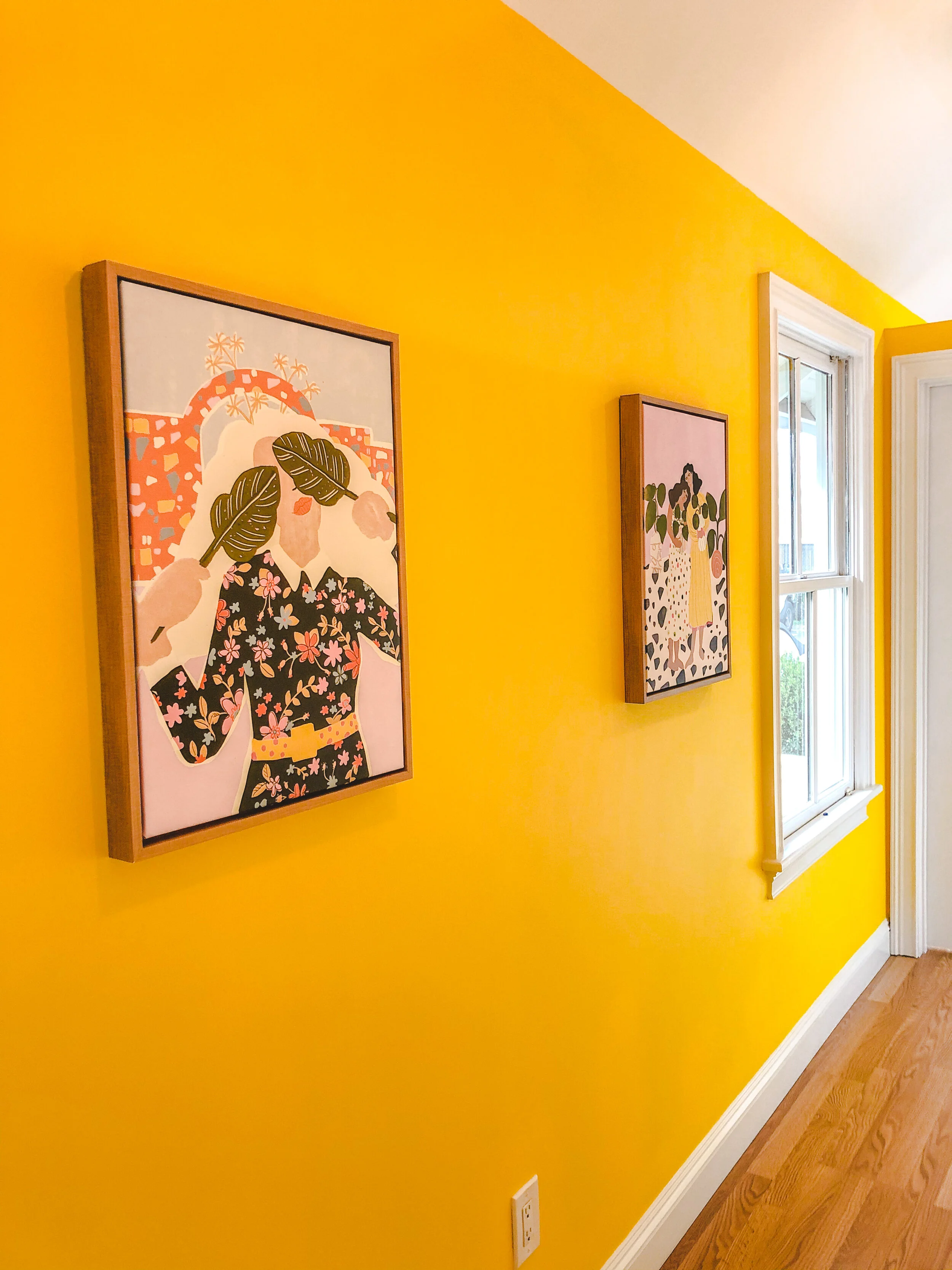 Yellow hallway with two framed artworks featuring colorful illustrations. One artwork depicts a figure with floral patterns and large leaves; the other shows a figure with polka dots and abstract shapes. Wooden floor and a window on the right.