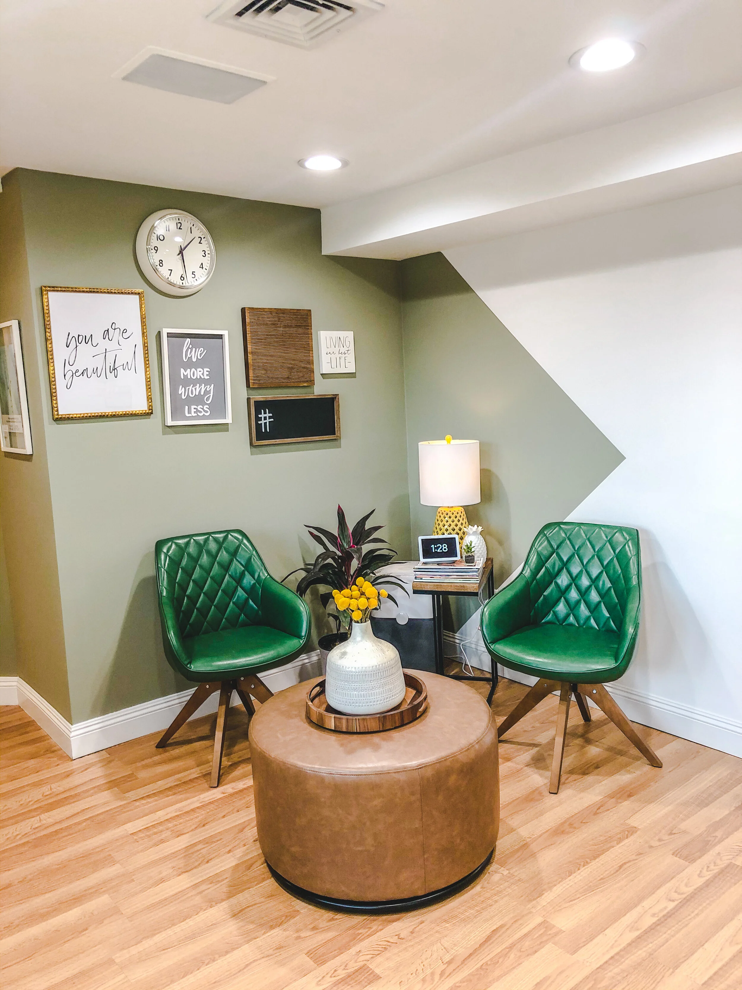 Small seating area with two green chairs and a round brown ottoman. A side table with a lamp and clock are nearby. Decorative wall art and a clock hang on a sage green wall. Wooden flooring is visible.