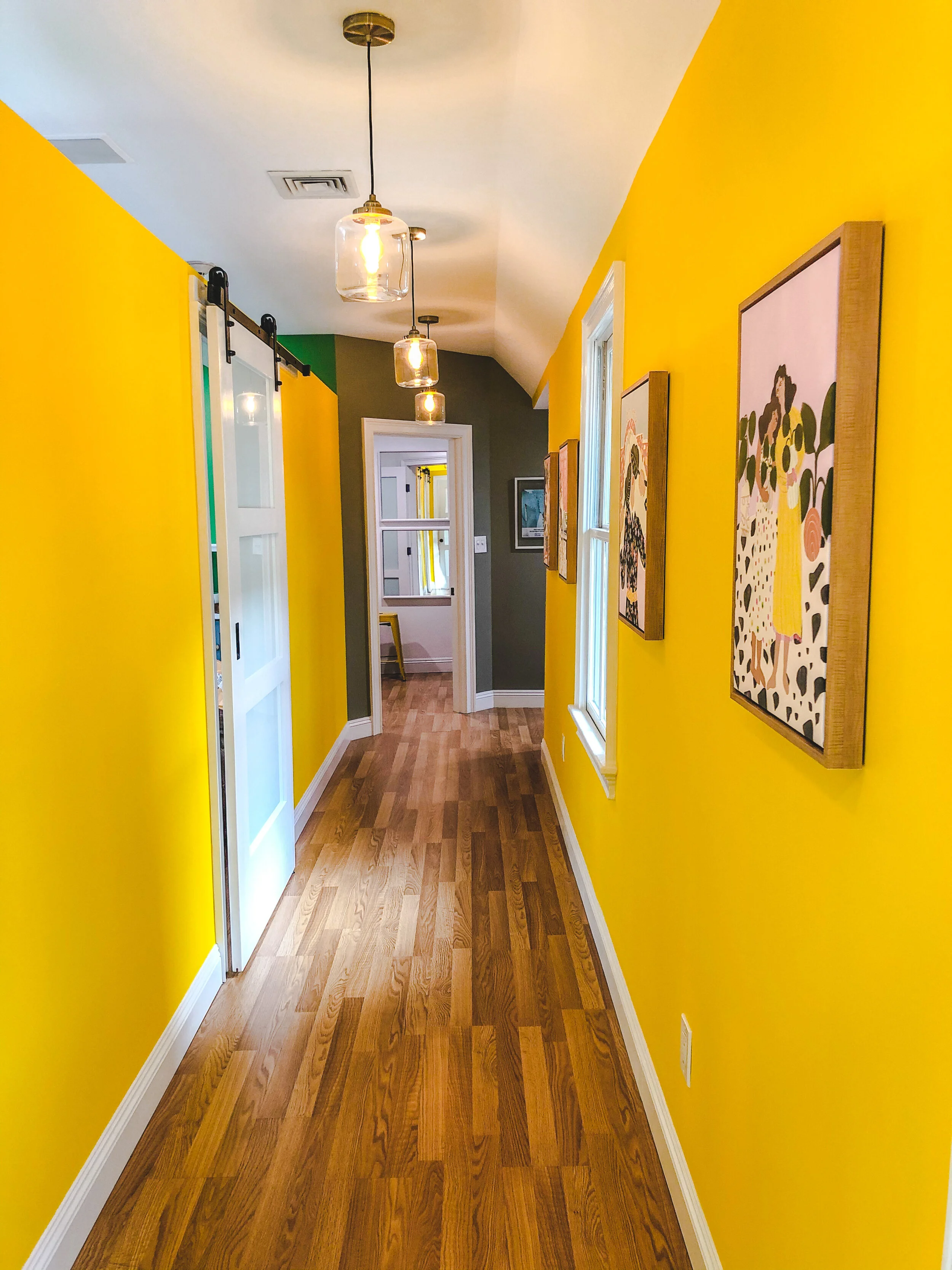 Vibrantly painted yellow hallway with wood flooring, featuring hanging pendant lights, wall art, and a sliding white barn door.