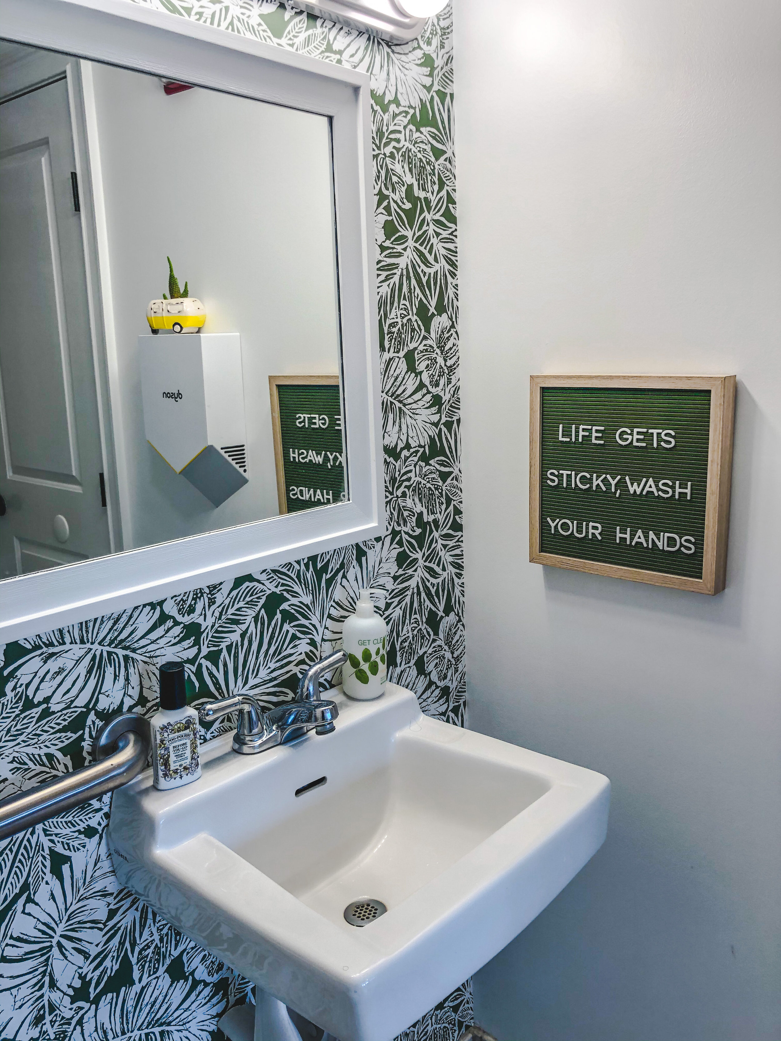 Bathroom interior with a white sink, tropical leaf-patterned wallpaper, a sign reading "Life gets sticky, wash your hands," and soap dispensers.