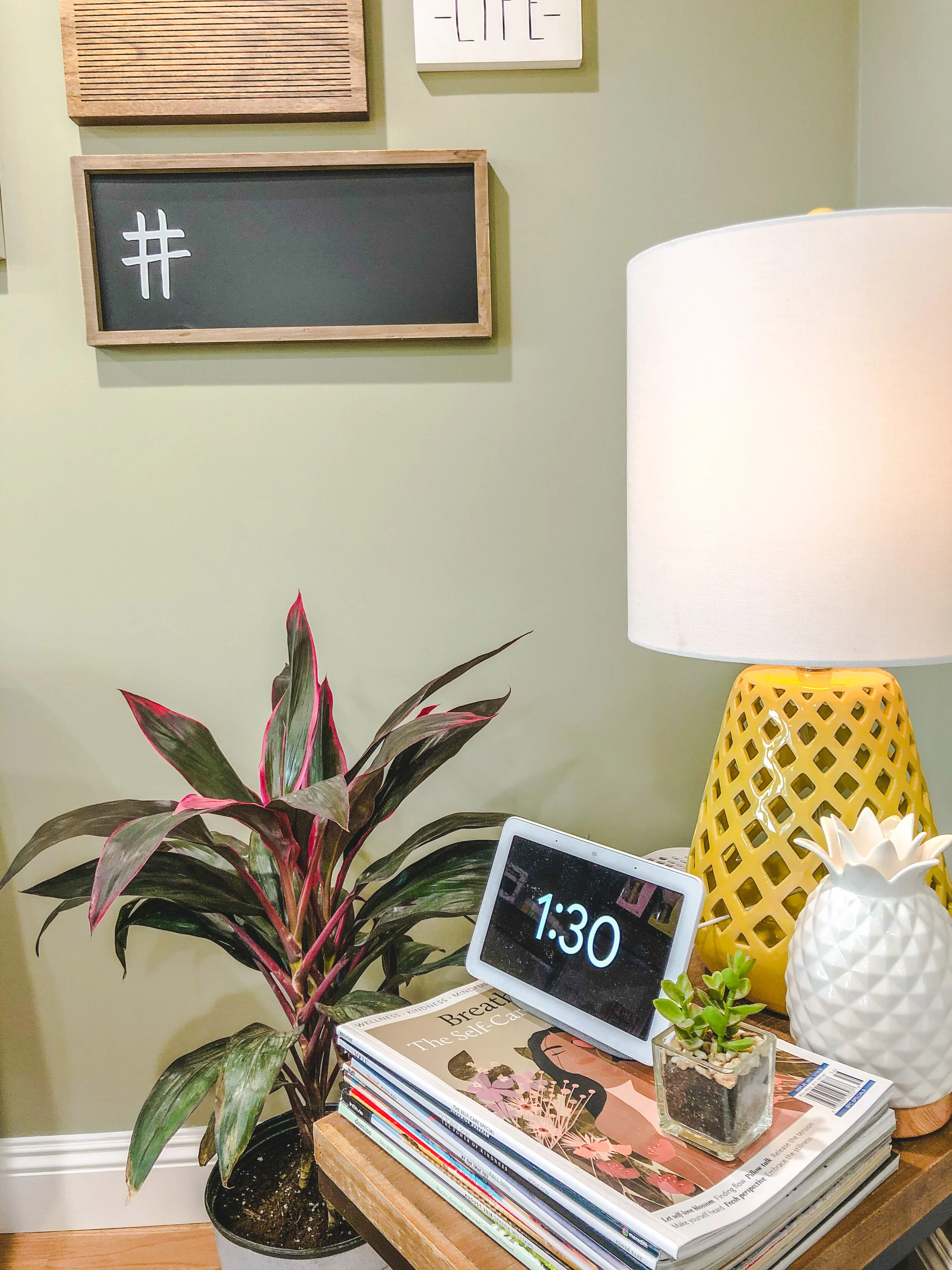 Home interior with a digital clock displaying 1:30, a potted houseplant, a stack of magazines, a yellow lattice lamp, a white pineapple-shaped object, and wall decor with a hashtag symbol on a wooden board.