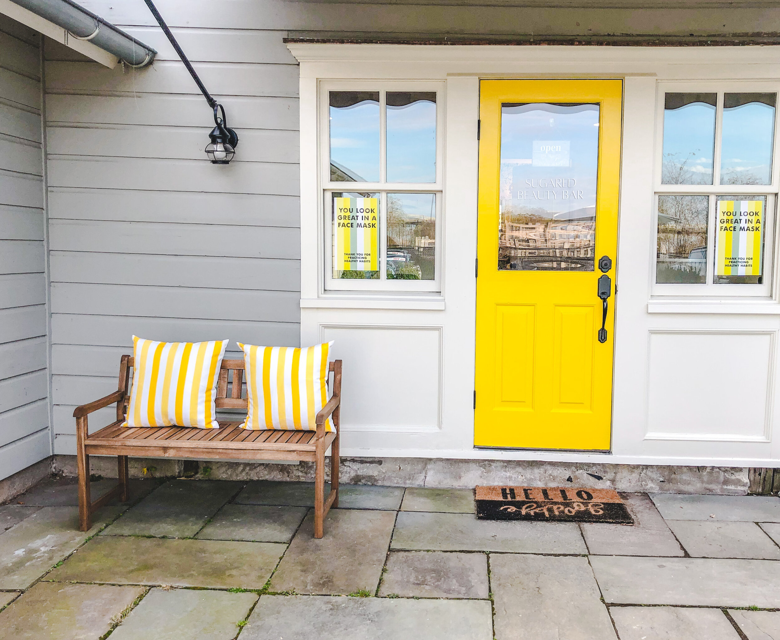 Yellow door with 'Sugared Beauty Bar' signage, flanked by windows displaying "You look great in a face mask" posters, wooden bench with yellow-striped pillows, and "Hello Gorgeous" doormat.