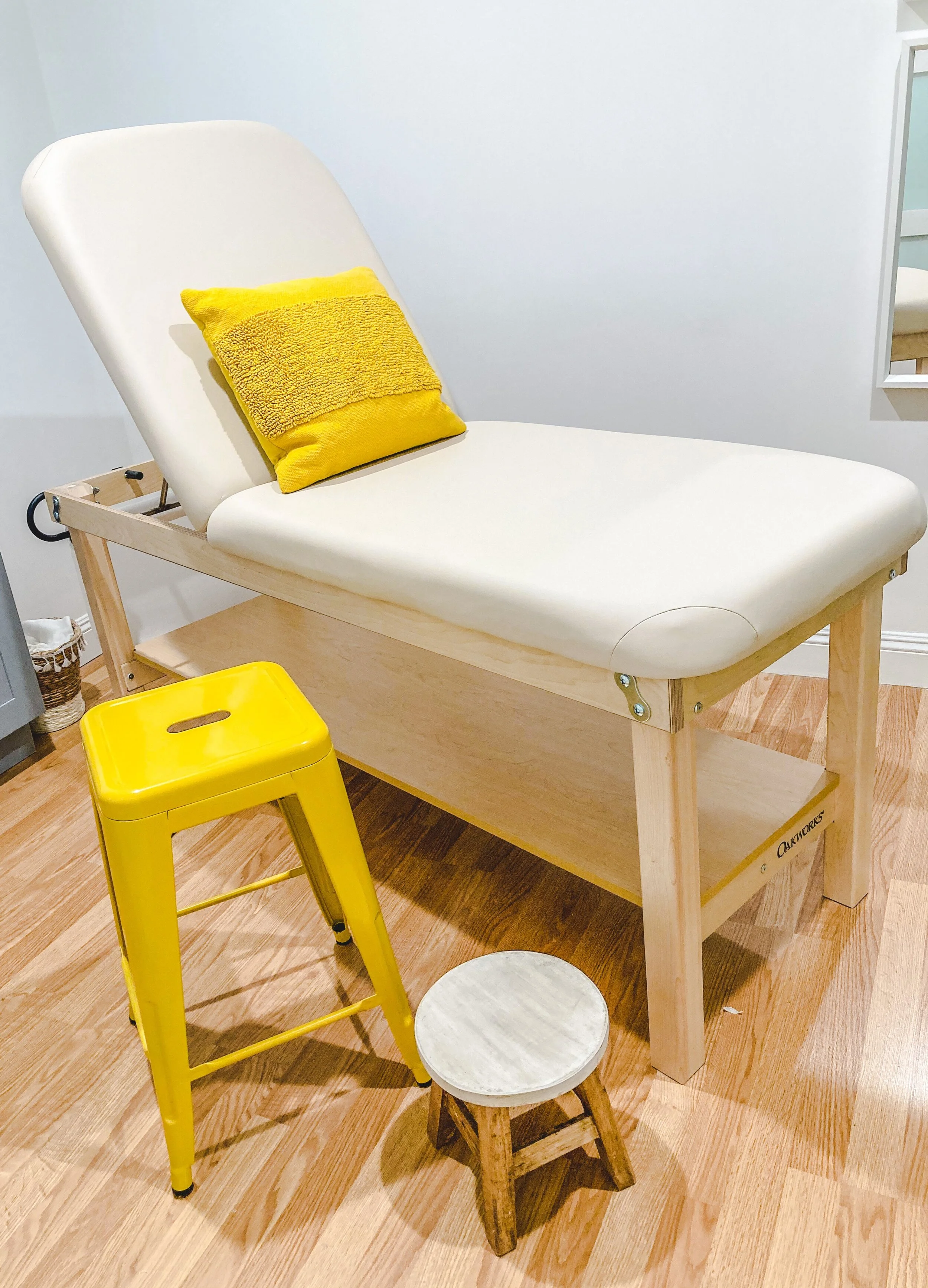 Modern massage or examination table with beige cushion, a yellow pillow, a bright yellow stool, and a small wooden stool on a wooden floor.