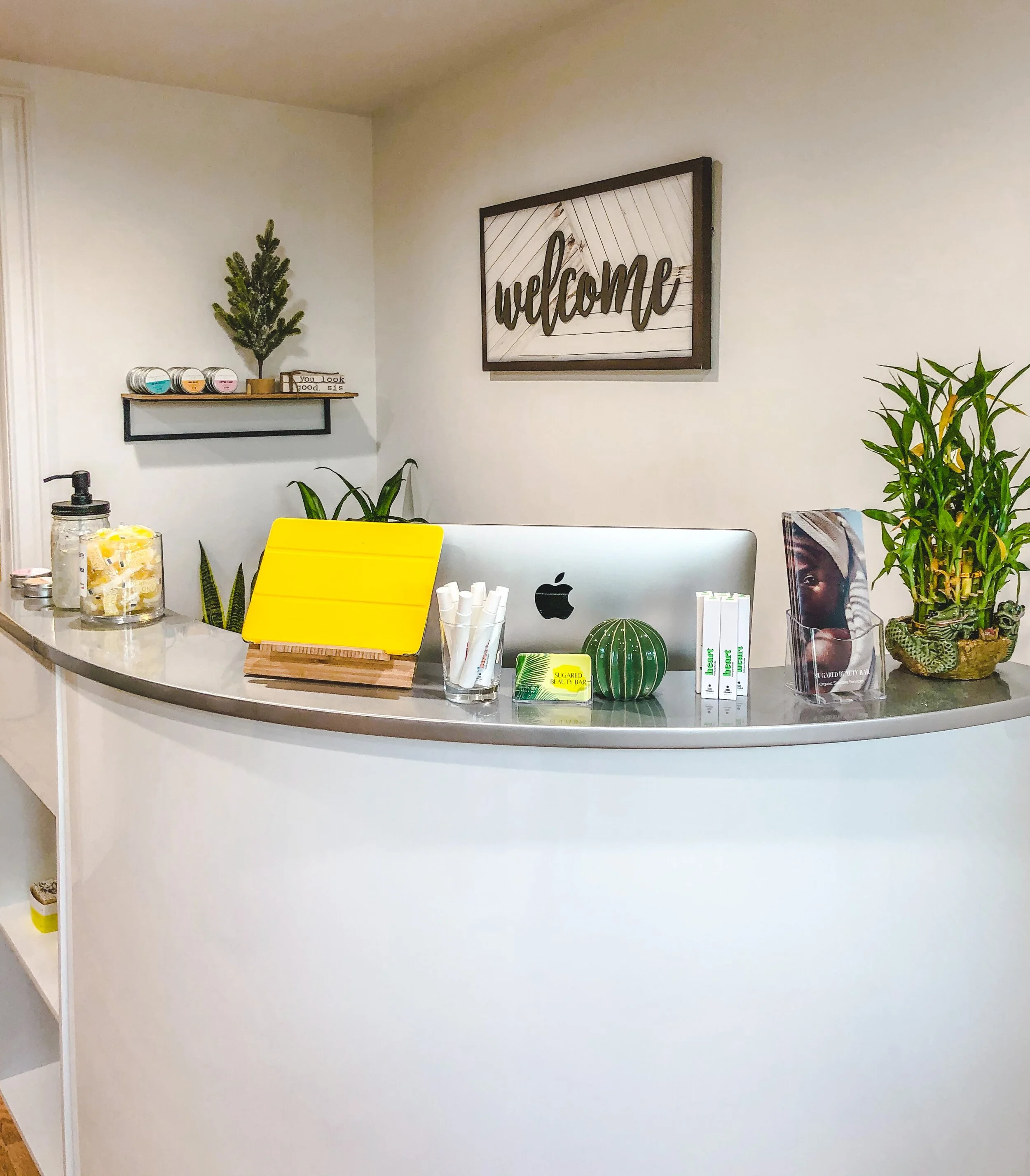Reception desk with an iMac, yellow tablet cover, bamboo plants, and welcome sign on the wall.