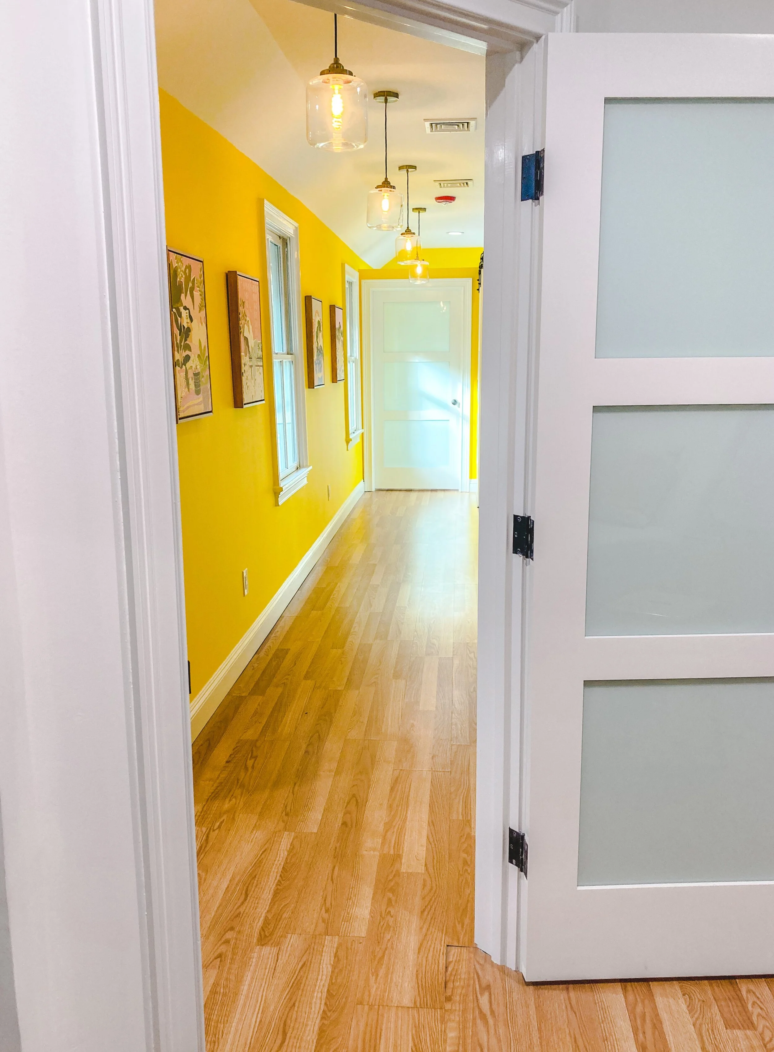 Bright hallway with yellow walls, wood flooring, pendant lights, framed artwork, and a white frosted glass door.