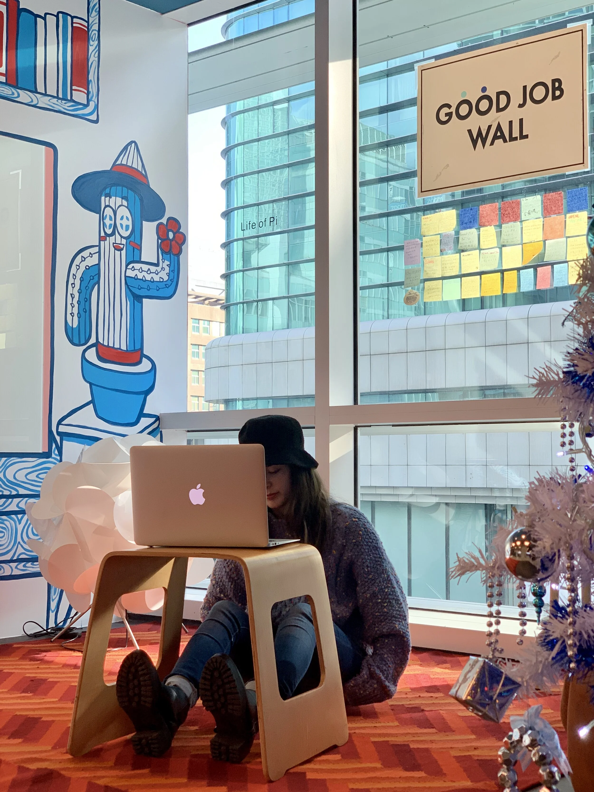 A woman sitting on the floor with legs extended, working on a MacBook at a small wooden table, wearing a black hat. The room has a colorful, patterned carpet, a decorated Christmas tree to the right, and large windows showing modern buildings outside