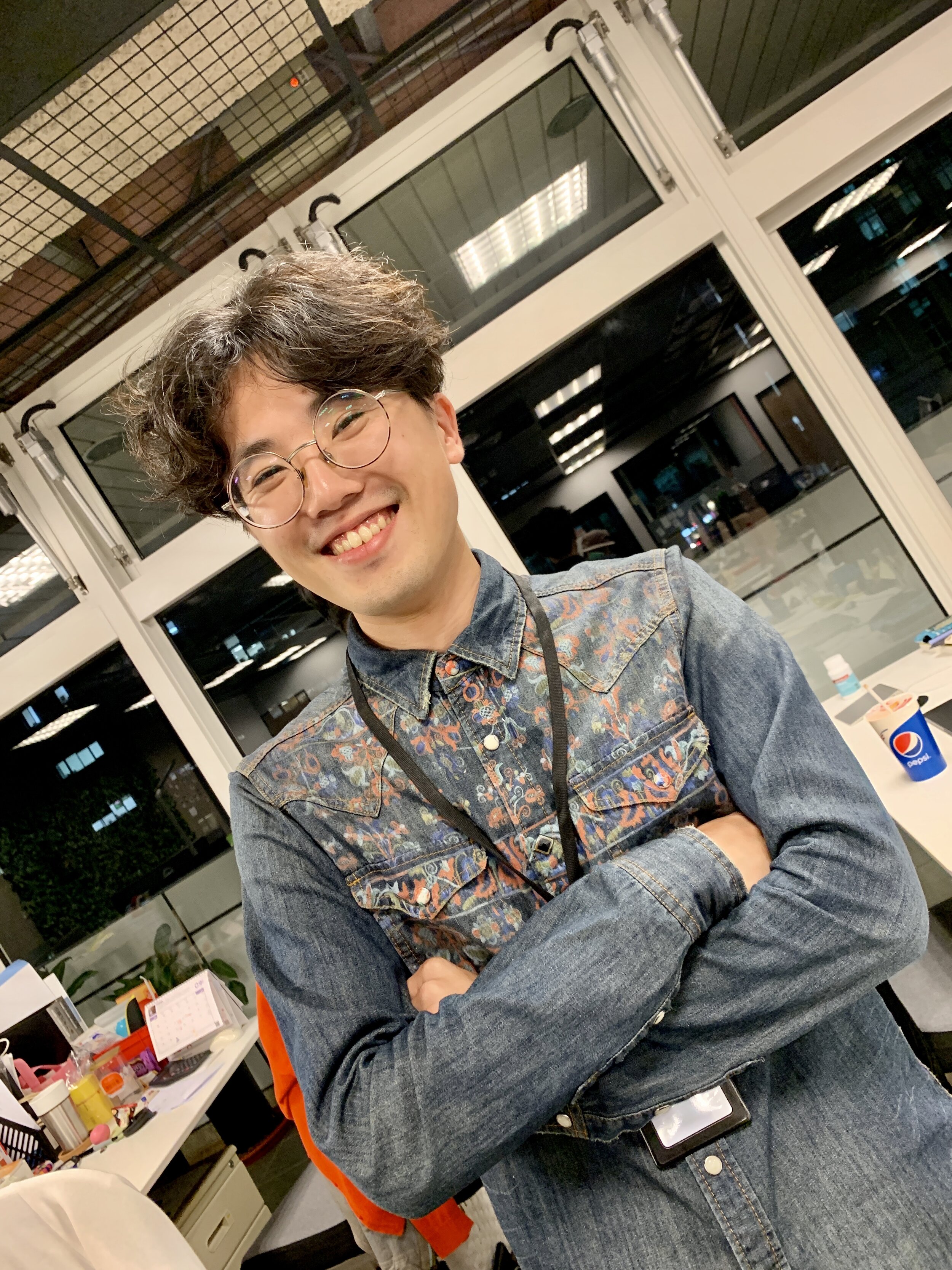 A young man with glasses, curly hair, wearing a denim jacket with embroidered details, smiling with arms crossed inside an office or workspace environment.
