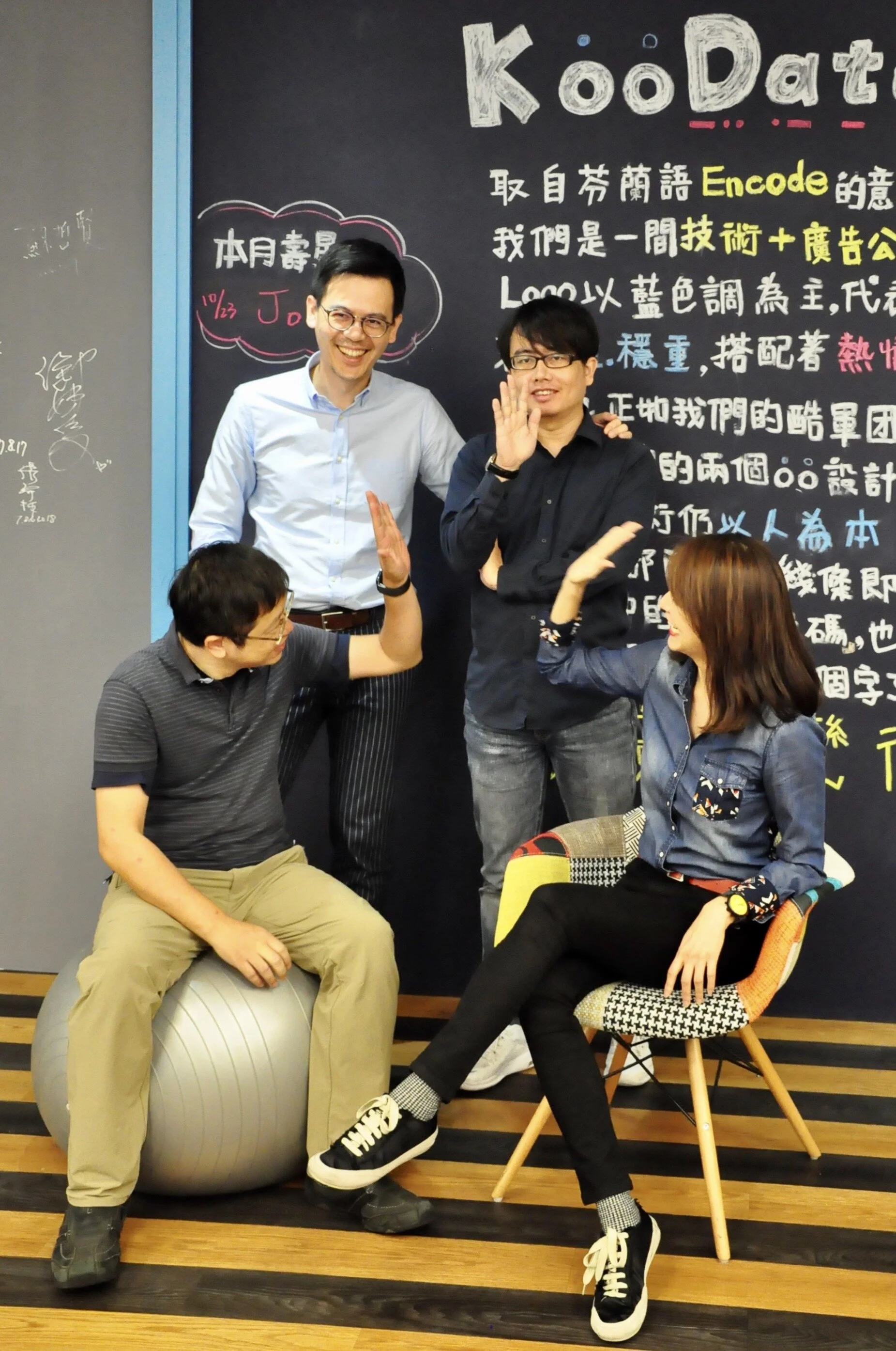 Four people in a casual setting, with three standing and two seated in front of a blackboard with handwritten text, some in Chinese characters and some in English, including the word 'KooDaat'. They are smiling, giving high-fives, and interacting pla
