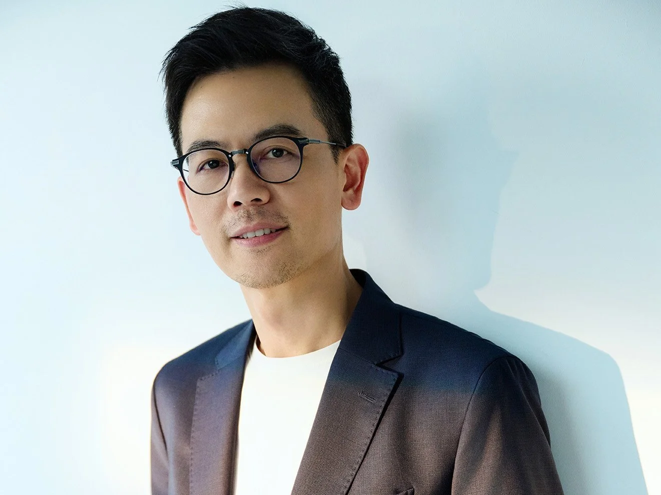 A young man with black hair and glasses wearing a dark blazer against a white background.