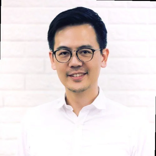 A young man with black hair and glasses, smiling, wearing a white shirt, standing against a white brick background.