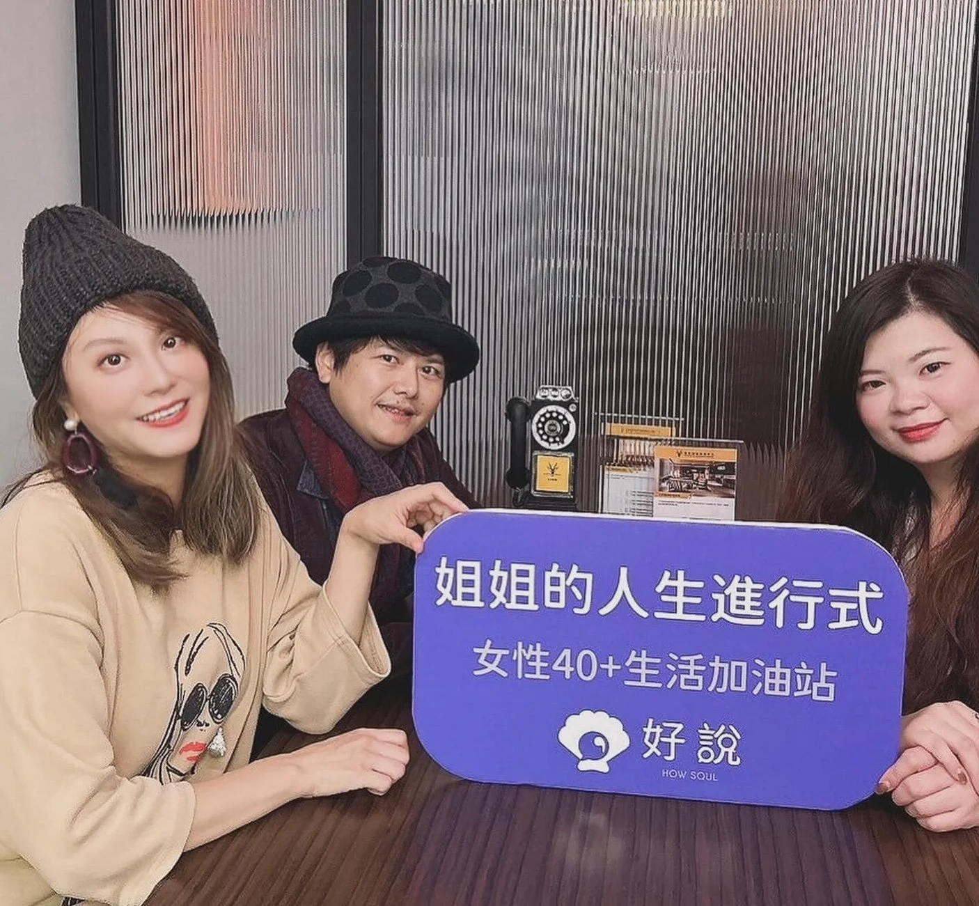 Three women sitting at a table holding a purple sign with Chinese text and a logo in a restaurant or cafe.
