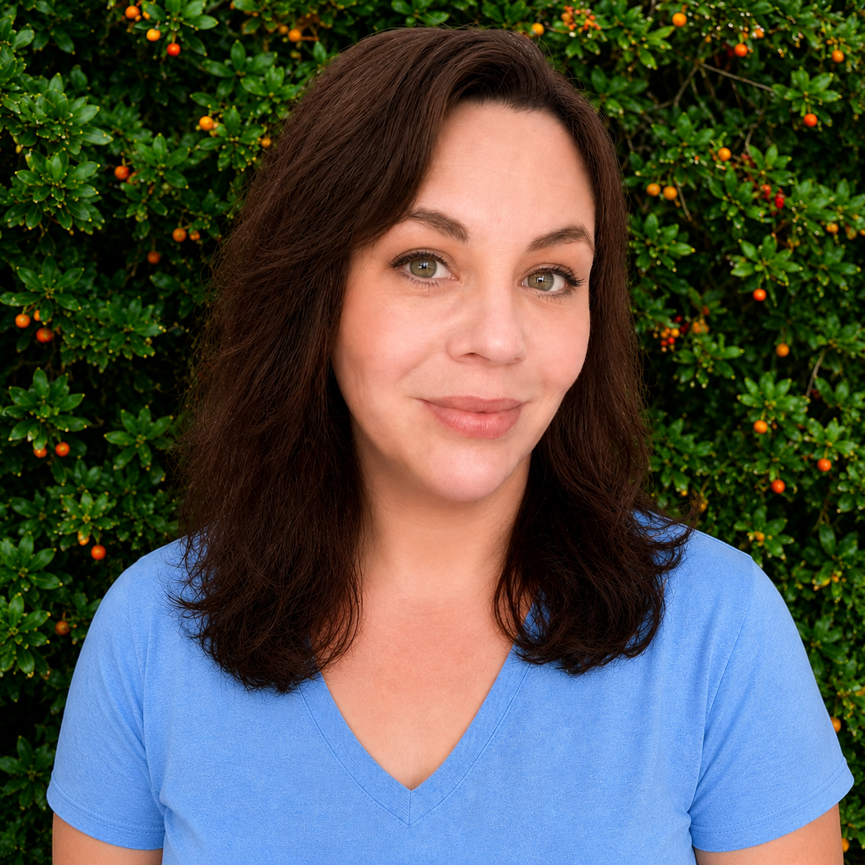 A woman with brown hair and green eyes smiling in front of a green shrub with small orange berries, wearing a light blue shirt.