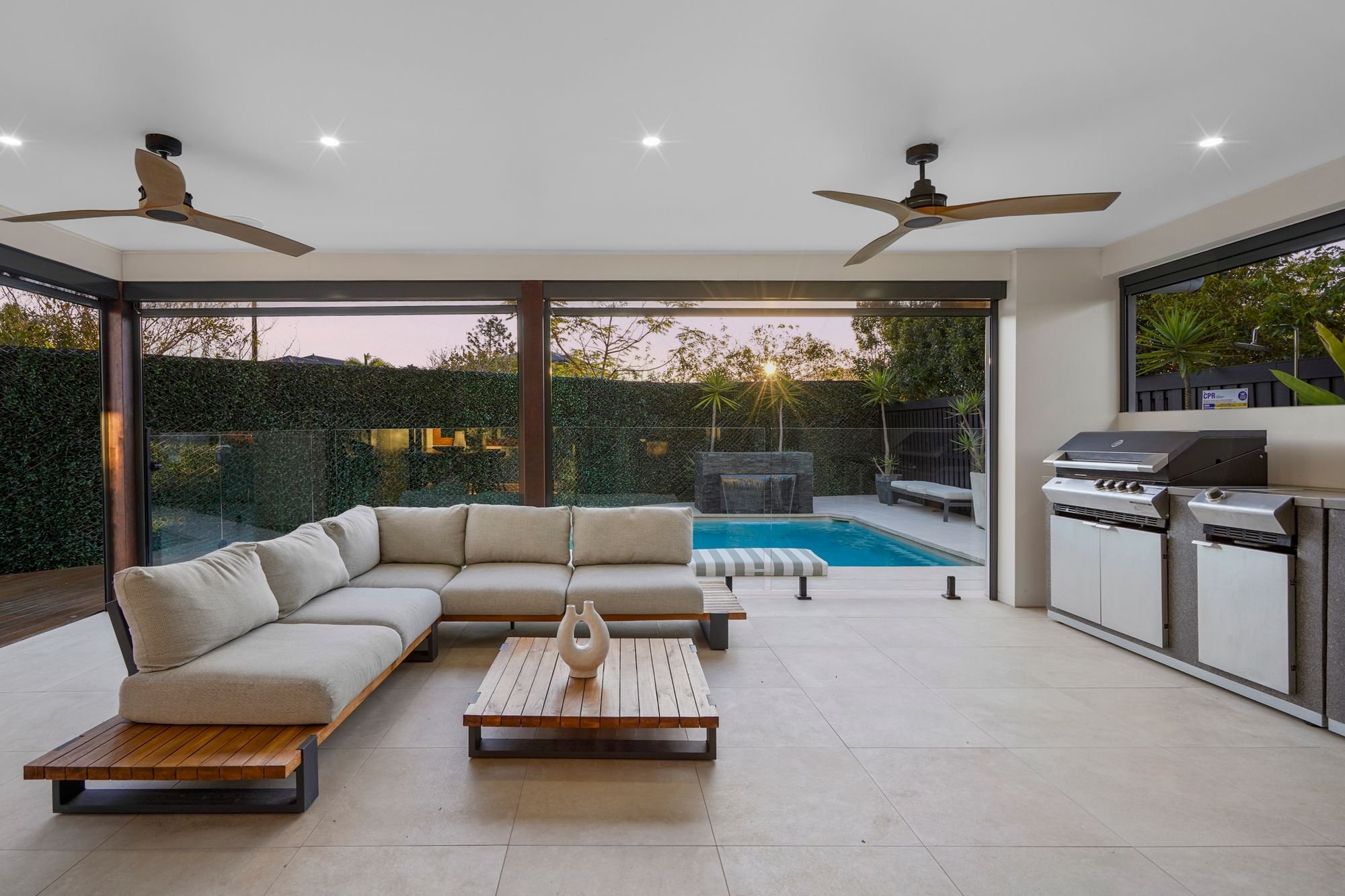 Indoor living area with large beige sectional sofa, wooden coffee table, and decorative vases. The room has sliding glass doors opening to an outdoor pool area with a waterfall feature and lawn, illuminated by sunset light. Ceiling fans and recessed lighting are also visible.