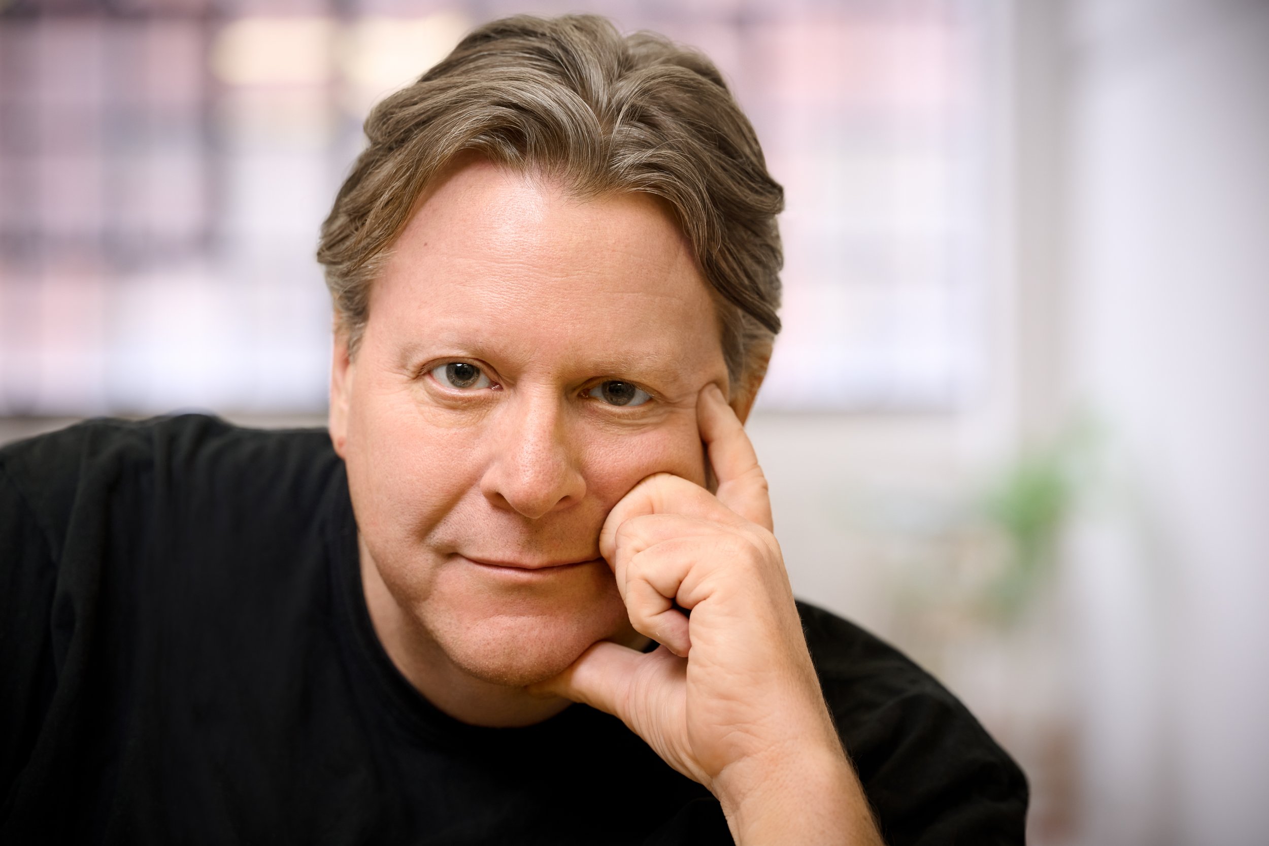 Close-up of a middle-aged man with brown hair, resting his face on his hand, looking directly at the camera with a slight smile, in an indoor setting with a blurred window in the background.