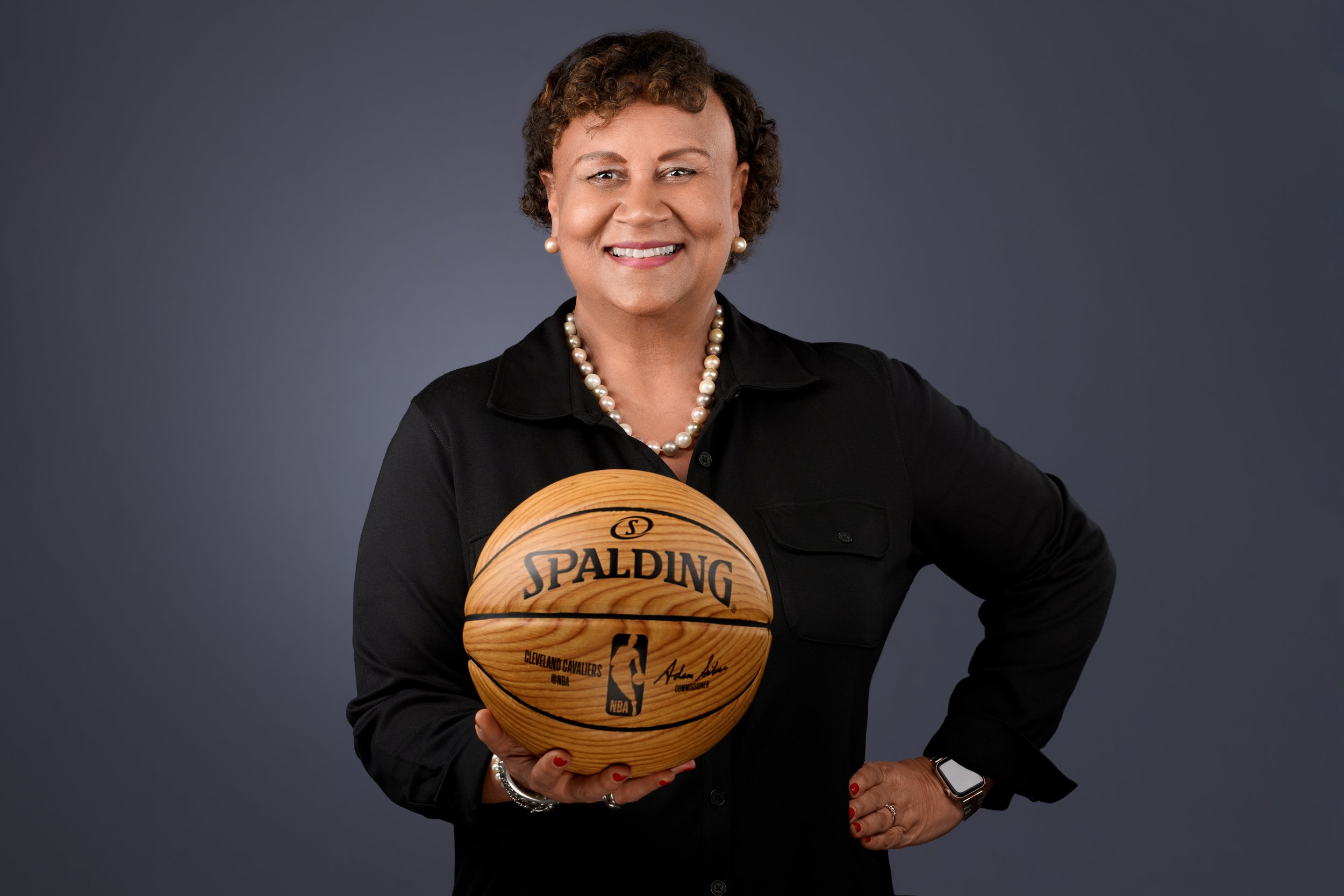 A woman with short curly hair, wearing a black shirt and pearl necklace, holding a signed Cleveland Cavaliers basketball, standing against a dark gray background.
