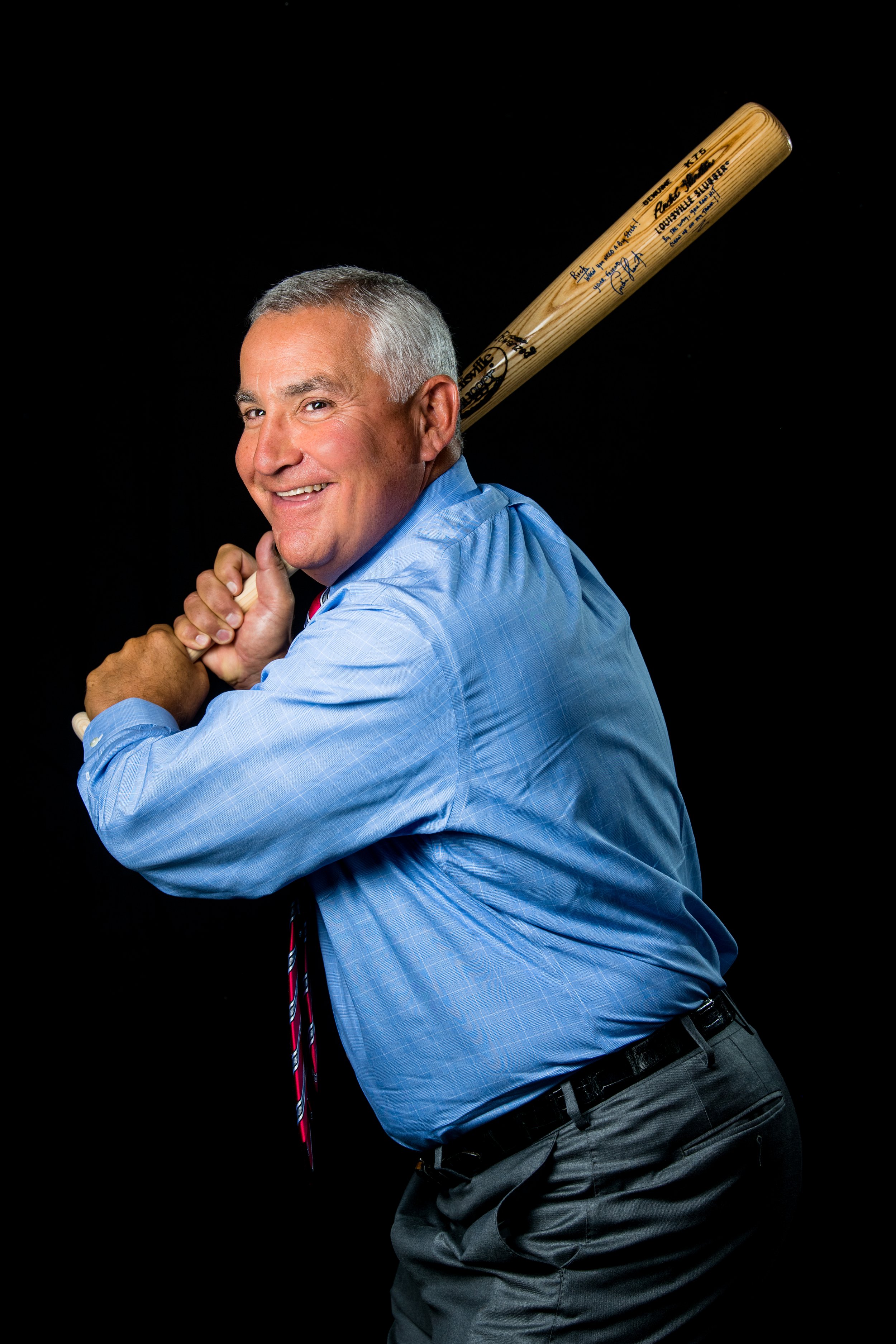 A man in a blue dress shirt and tie holding a baseball bat over his shoulder, smiling against a black background.