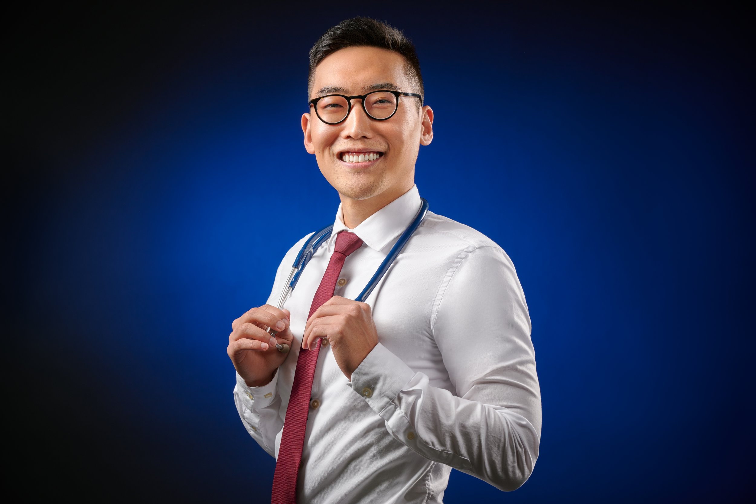 A smiling male doctor wearing glasses, a white shirt, a maroon tie, and a stethoscope around his neck, on a dark blue background.