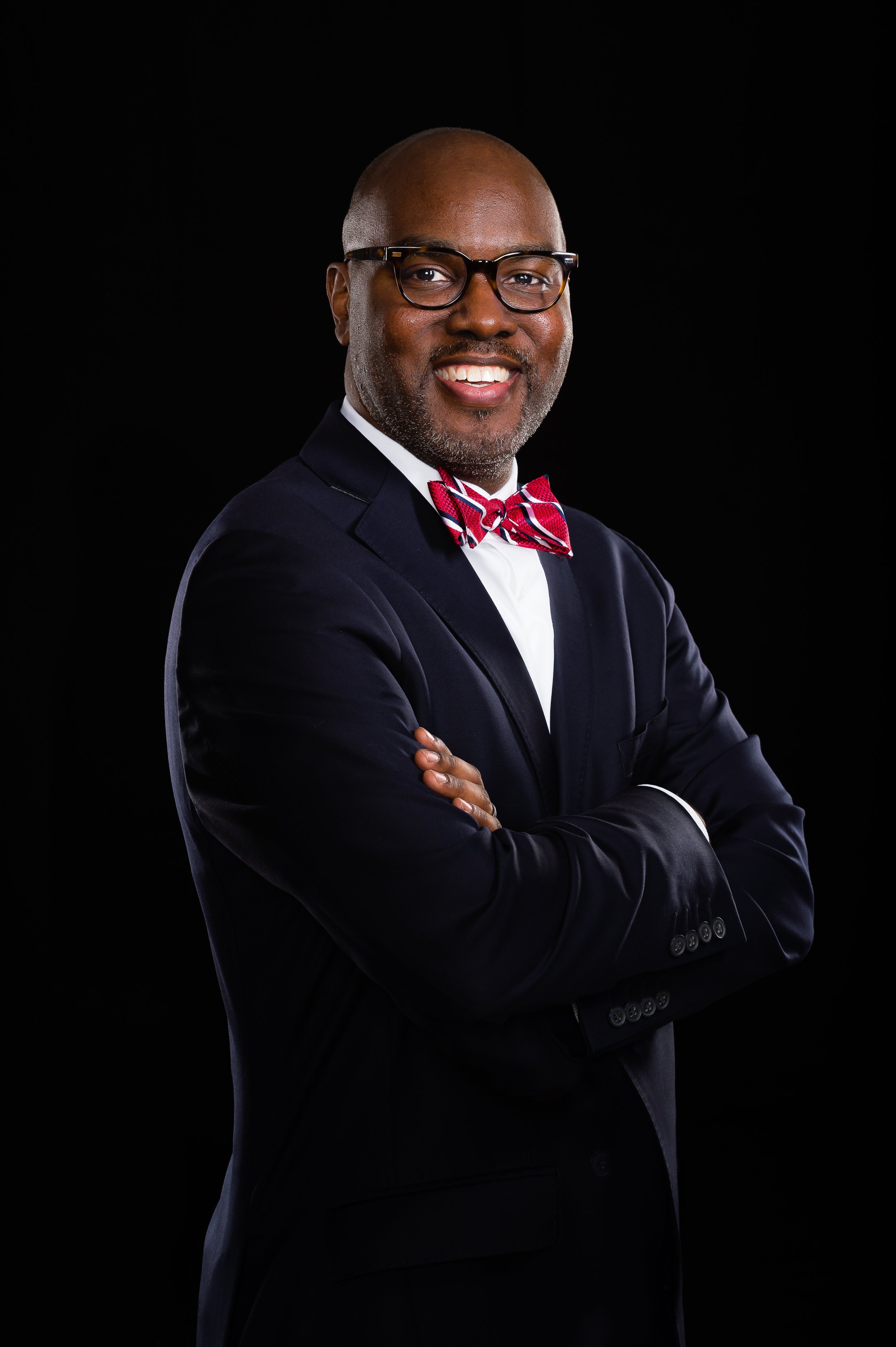 A smiling man wearing glasses, a dark suit, a white shirt, and a red patterned bow tie posing with arms crossed against a black background.