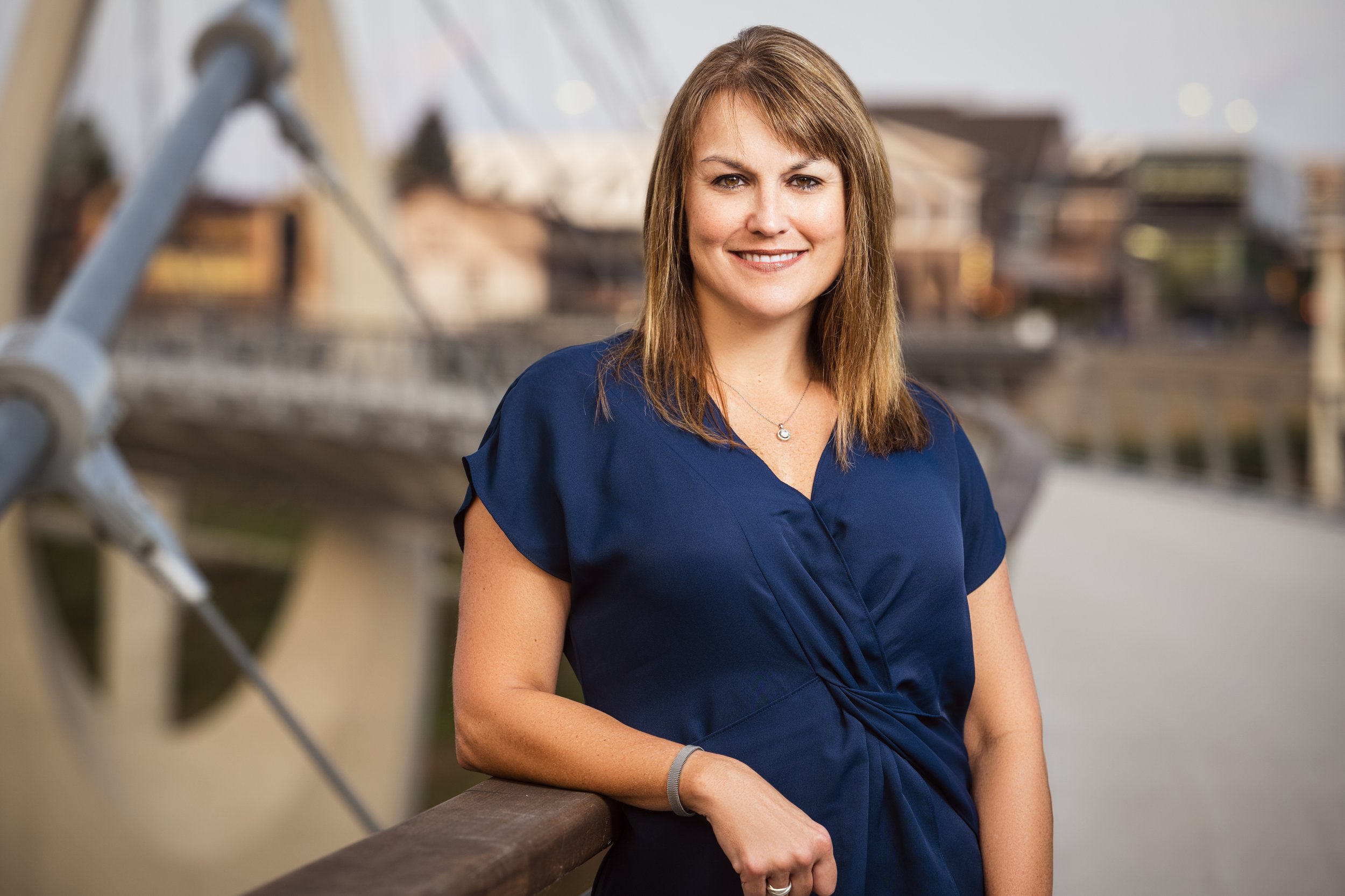 A woman with light brown hair wearing a blue dress, standing outdoors by a railing near a body of water with a blurred background of buildings and boats.