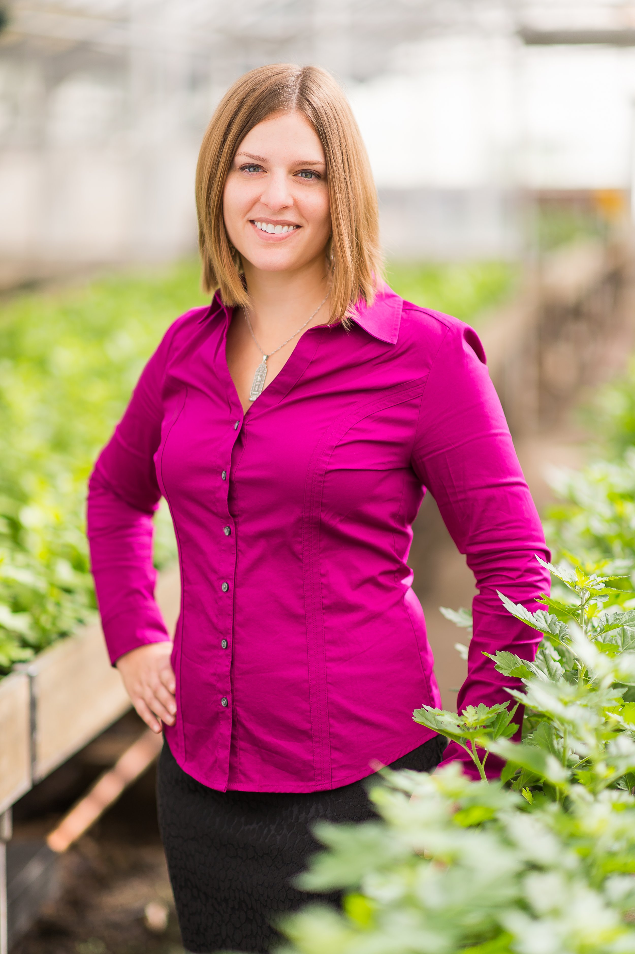 A woman with shoulder-length blonde hair, wearing a bright pink button-up shirt, standing in a greenhouse surrounded by green plants, smiling at the camera.