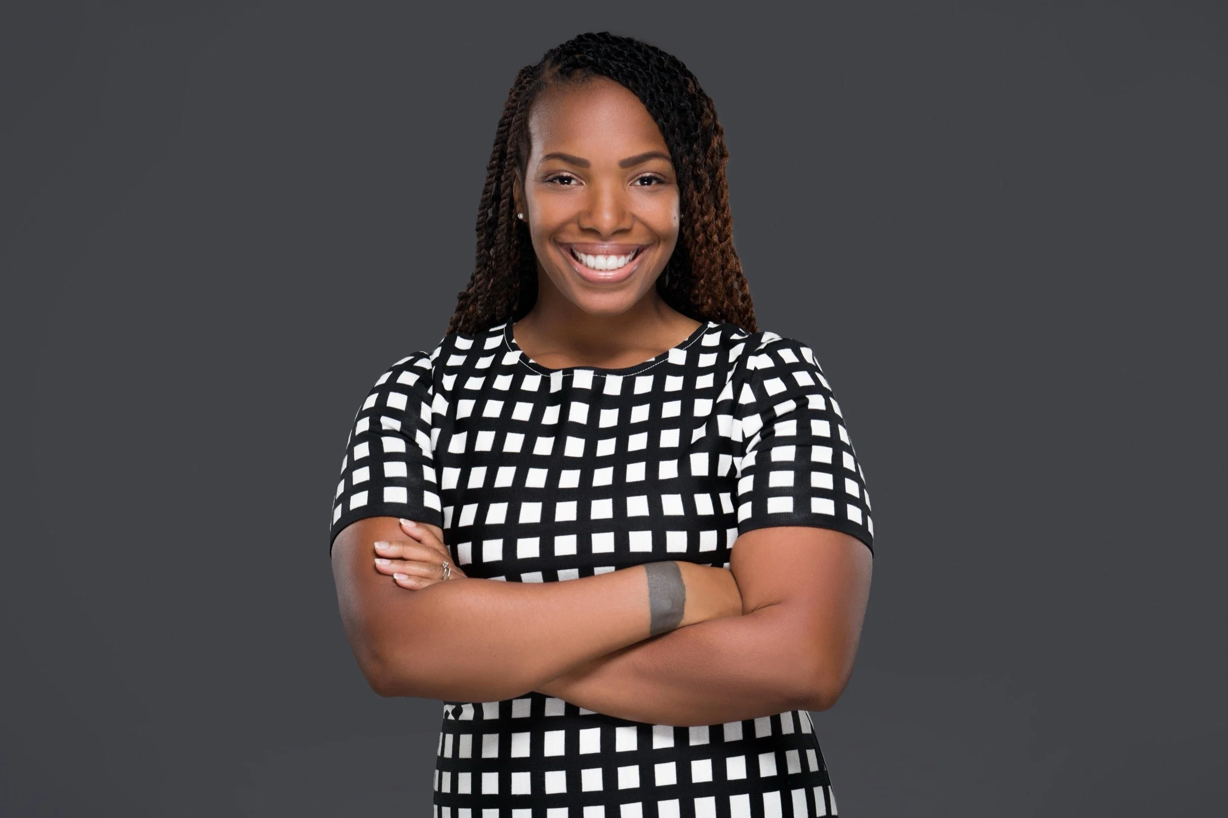 A smiling woman with crossed arms, wearing a black and white checkered dress, standing against a dark gray background.
