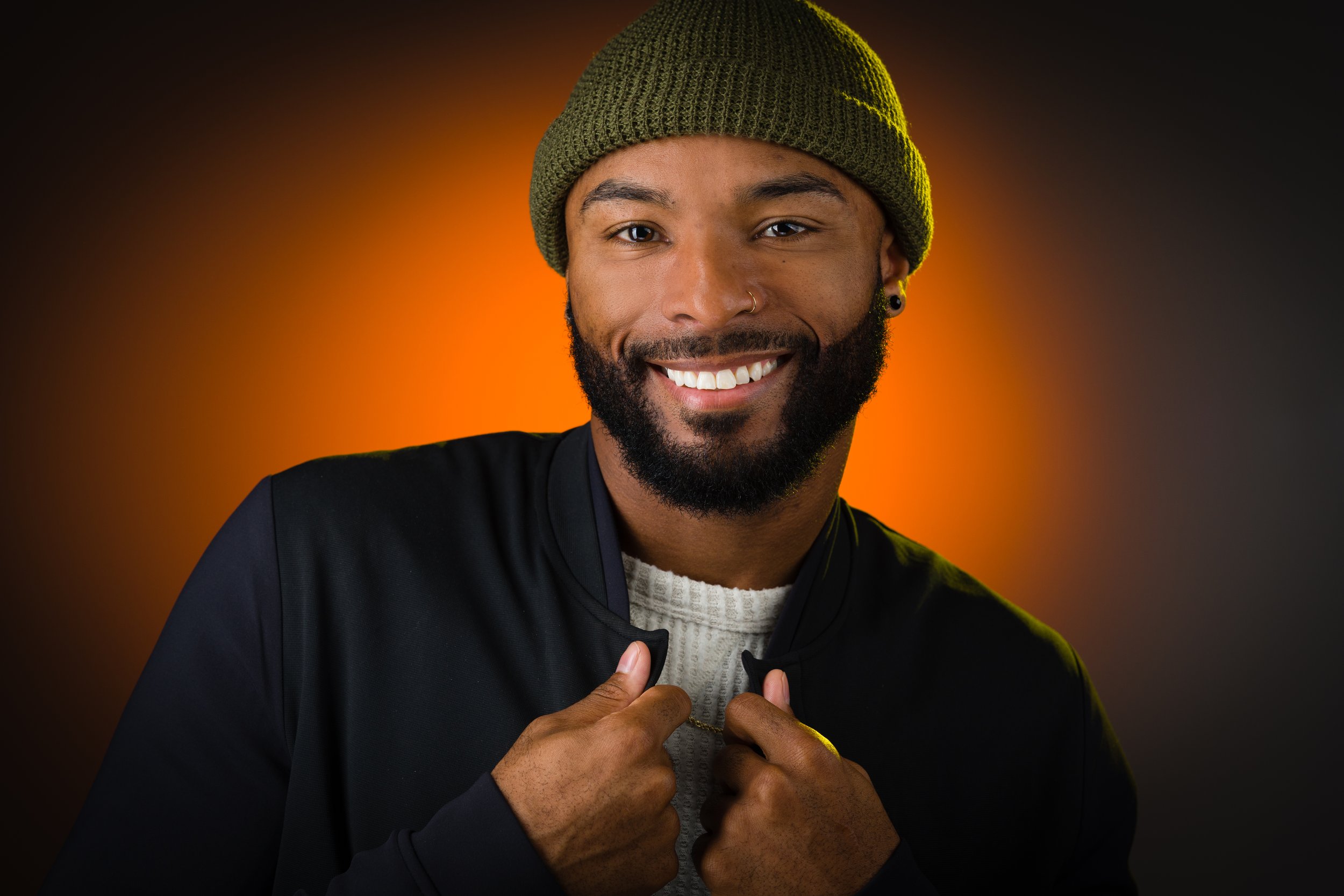 Smiling man wearing a green beanie, black jacket, and white sweater, holding the collar of his jacket with both hands, against an orange and black gradient background.